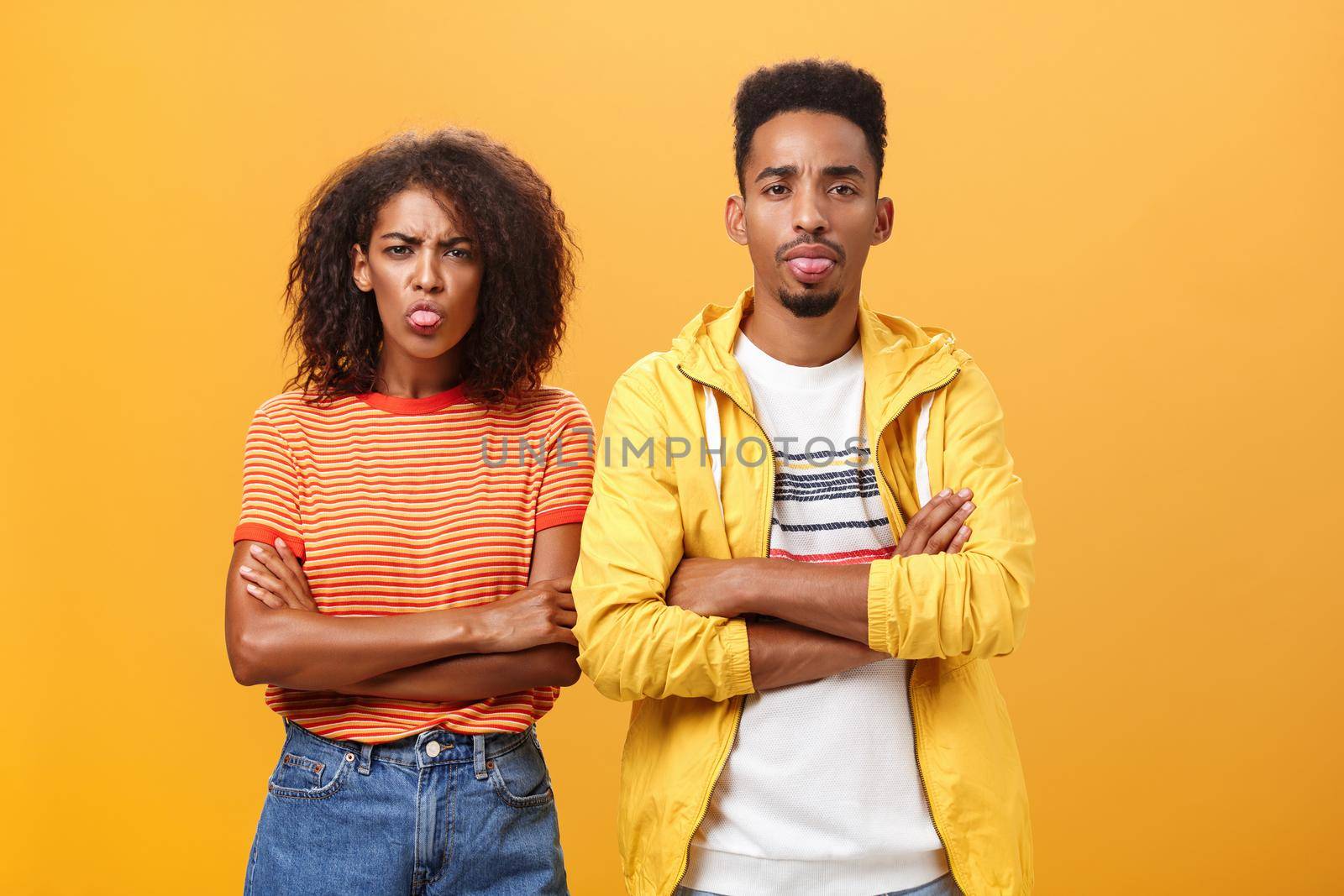 Indoor shot of african american siblings being displeased and annoyed showing bad tempber behaving childish sticking out tongue standing together with crossed hands on chest over orange background by Benzoix