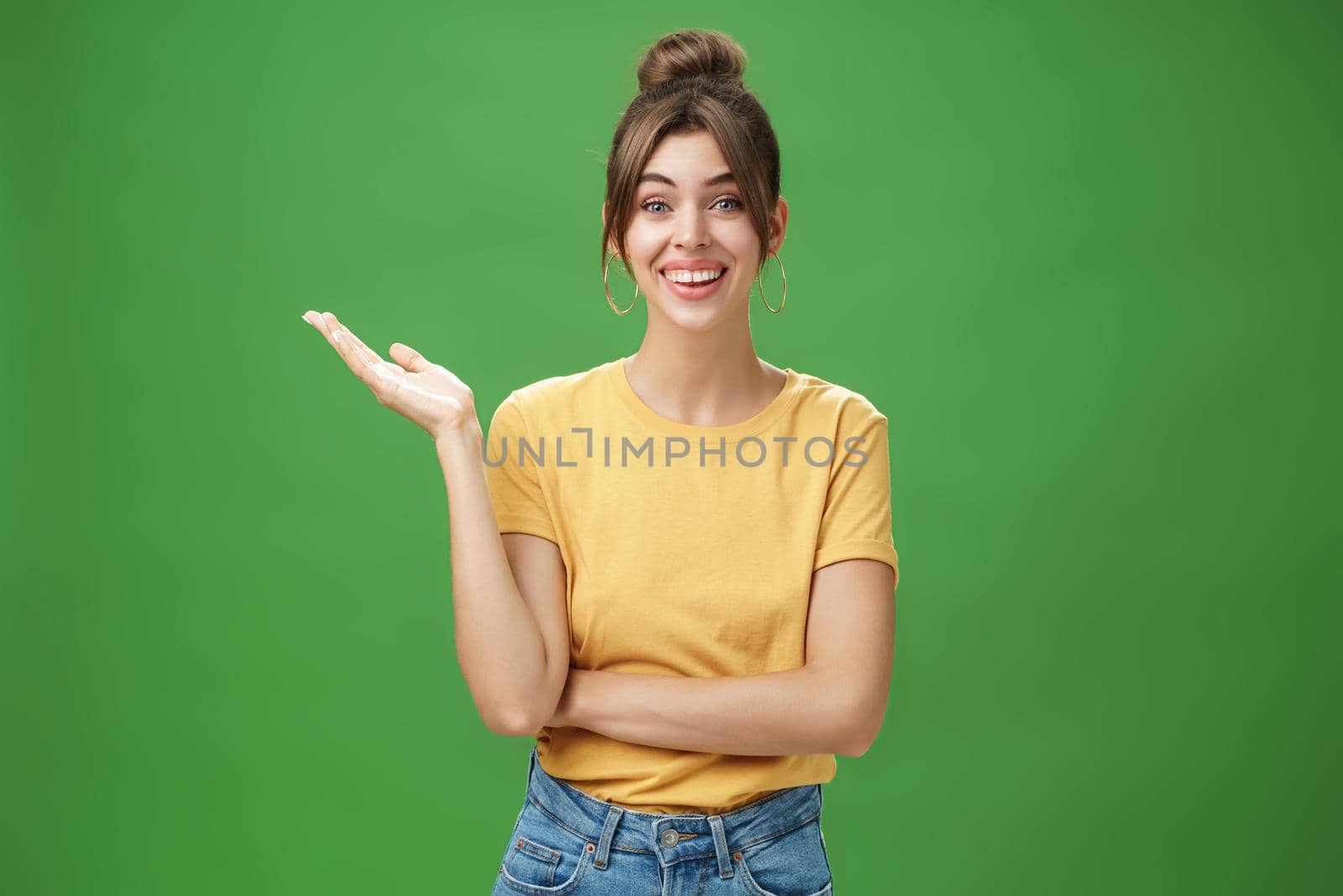 Wondered and carefree young friendly woman with combed hair cute gapped teeth in yellow t-shirt raising one hand in surprise reacting to amazing news smiling broadly at camera over green wall. Lifestyle.