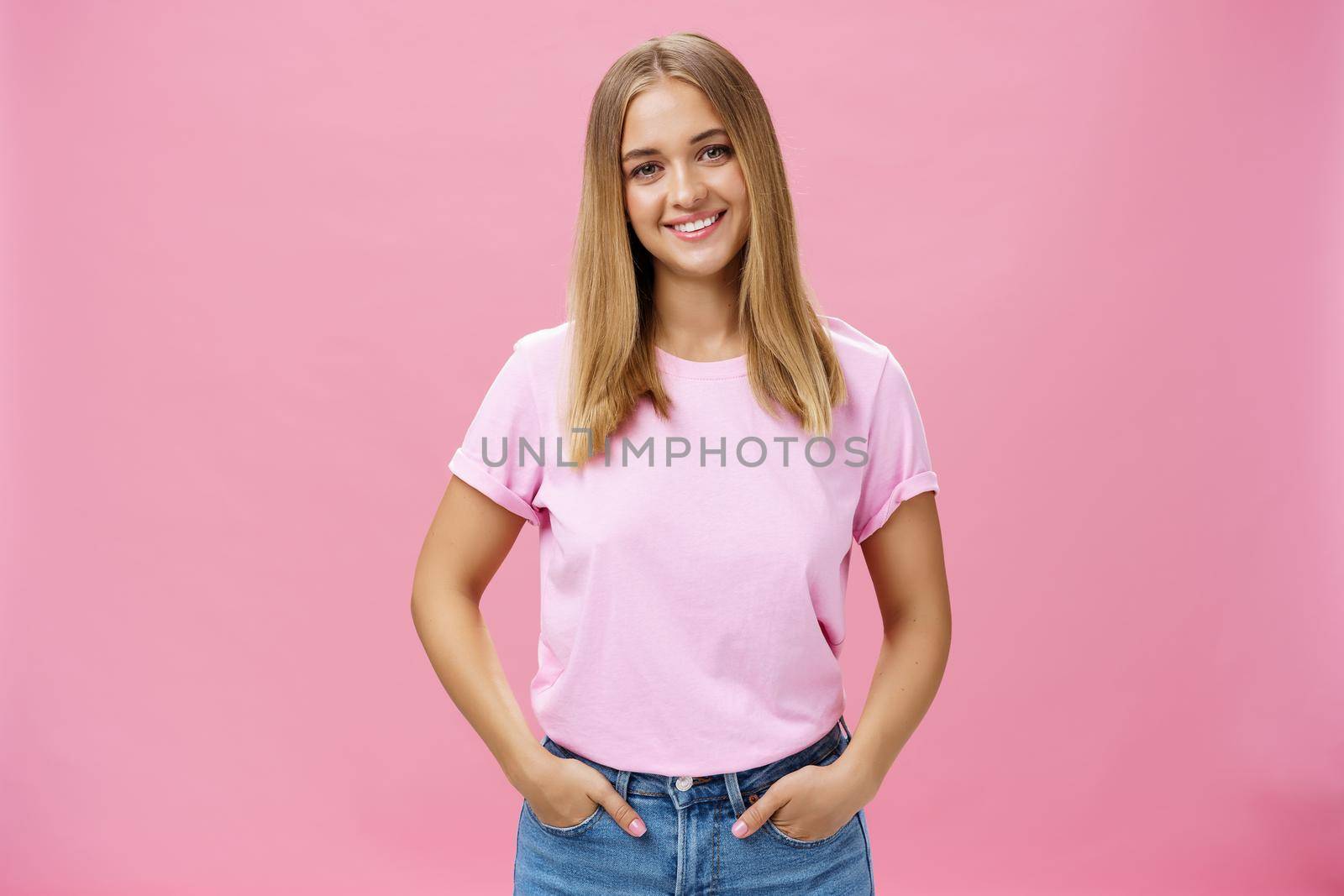 Charming pleasant woman with chubby face and fair hair in casual t-shirt smiling broadly holding hands in pockets being nice and friendly gazing carefree and calm at camera over pink background. Lifestyle.