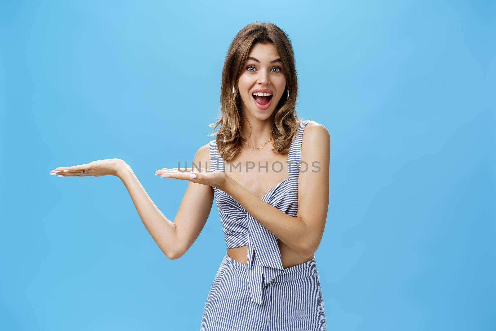 Woman presenting product over copy space. Good-looking charismatic adult tall woman with chestnut hair pointing at left side of wall and smiling broadly as if advertising against blue background.