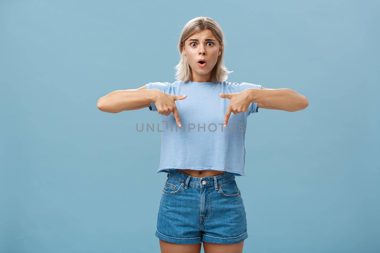 Studio shot of worried caucasian young woman feeling nervous her shoes do not fit frowning asking advice nervously pointing down shocked while frowning and standing over blue background by Benzoix
