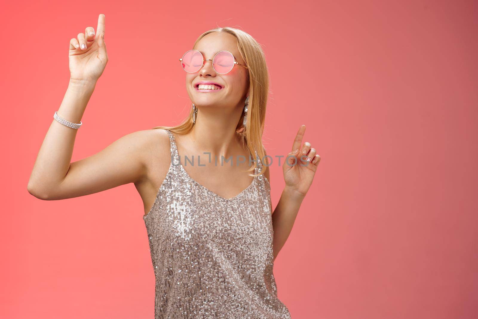 Delighted carefree attractive stylish millennial blond woman celebrating partying having fun wear sunglasses trendy silver dress dancing closed eyes broad smile waving hands up, red background.