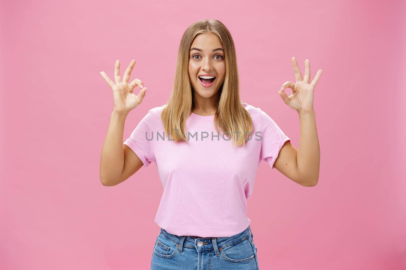 Portrait of enthusiastic attractive caucasian girl in trendy t-shirt and jeans showing okay or confirm gesture with amused broad smile standing pleased over pink background reacting to excellent news. Lifestyle.