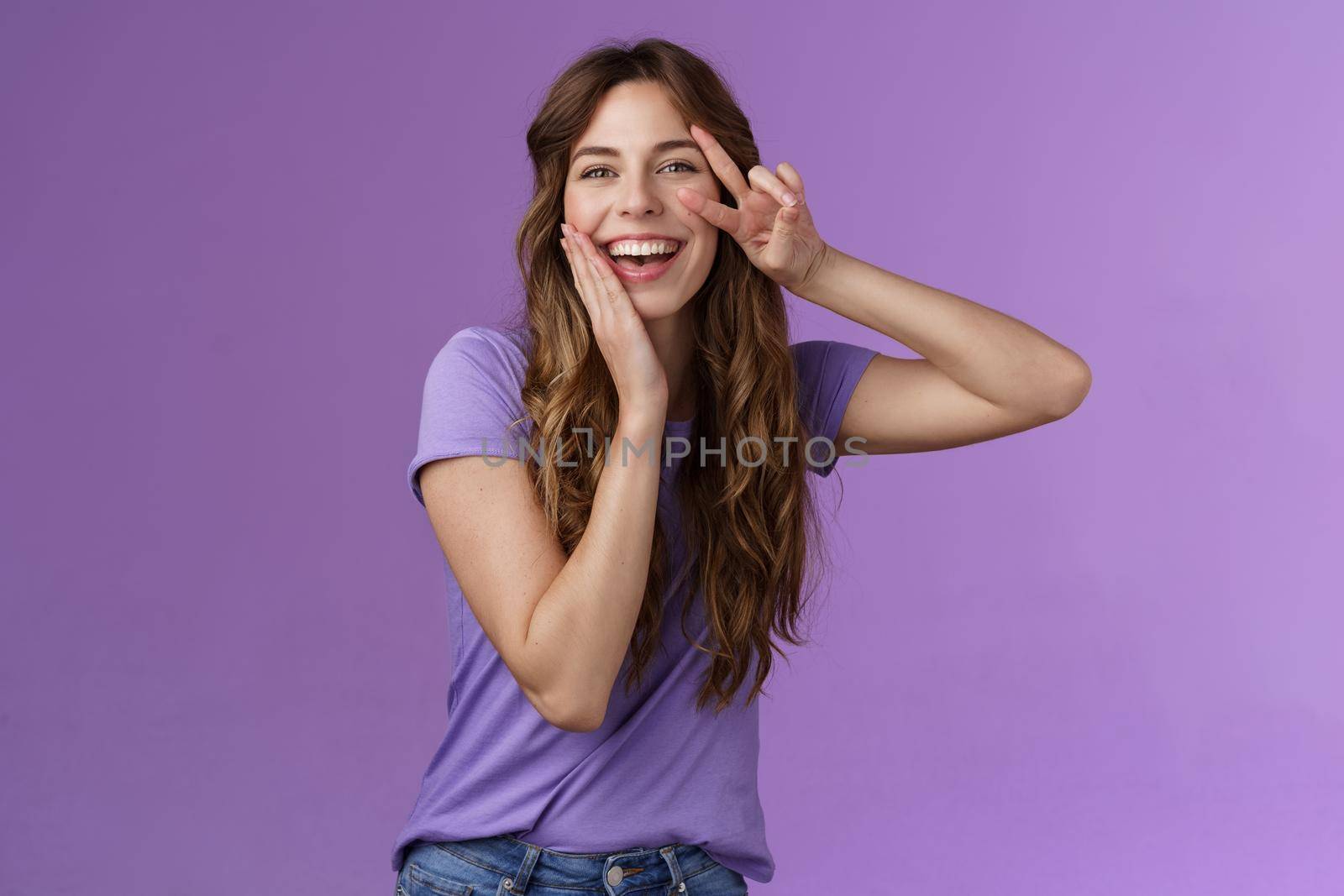 Adorable lovely funny cute woman curly brown hairstyle tilt head touch cheek blishing silly flirty smiling camera show peace victory sign express positive joyful attitude stand purple background. Lifestyle.