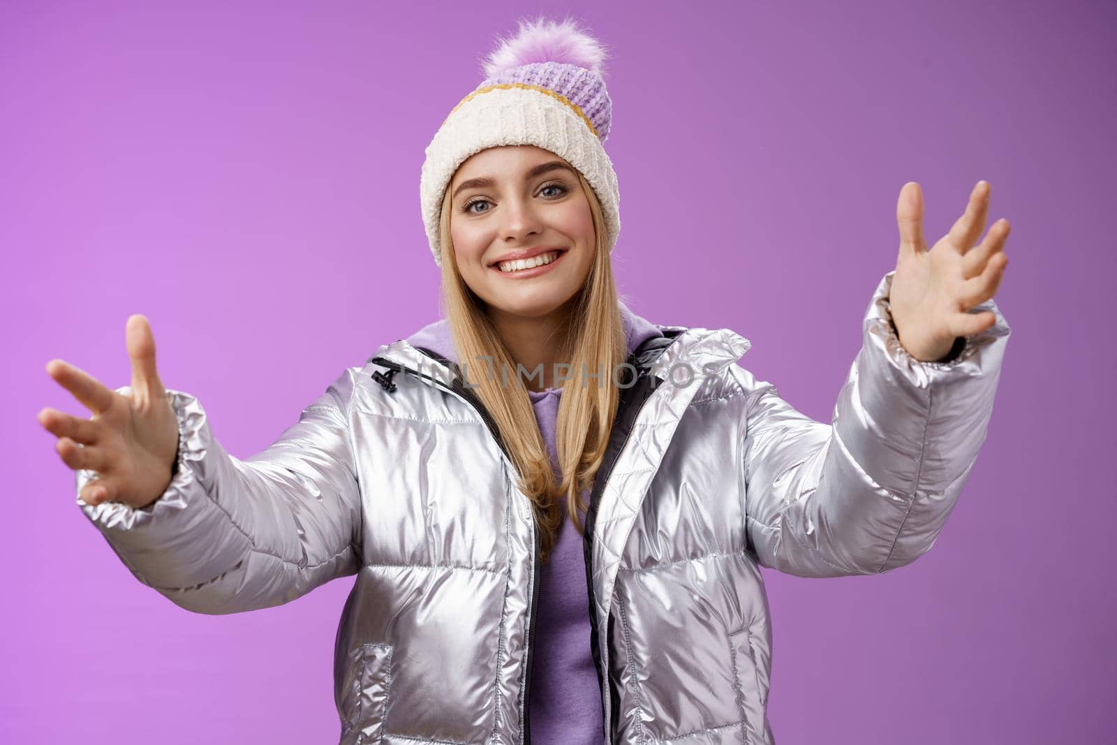 Friendly charming smiling european girl vacation wearing silver warm glittering jacket hat extend arms waiting girlfriend fall arms embrace loving person standing happily purple background.