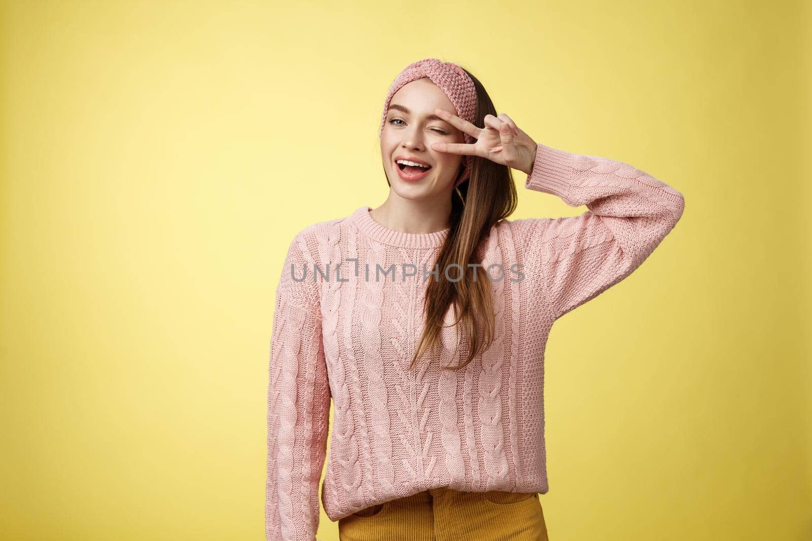 Cheerful happy glamour young european woman in pink knitted sweater, wearing headband, winking flirty and cute, showing victory or pease sign over eye, feeling excited and joyful over yellow wall.