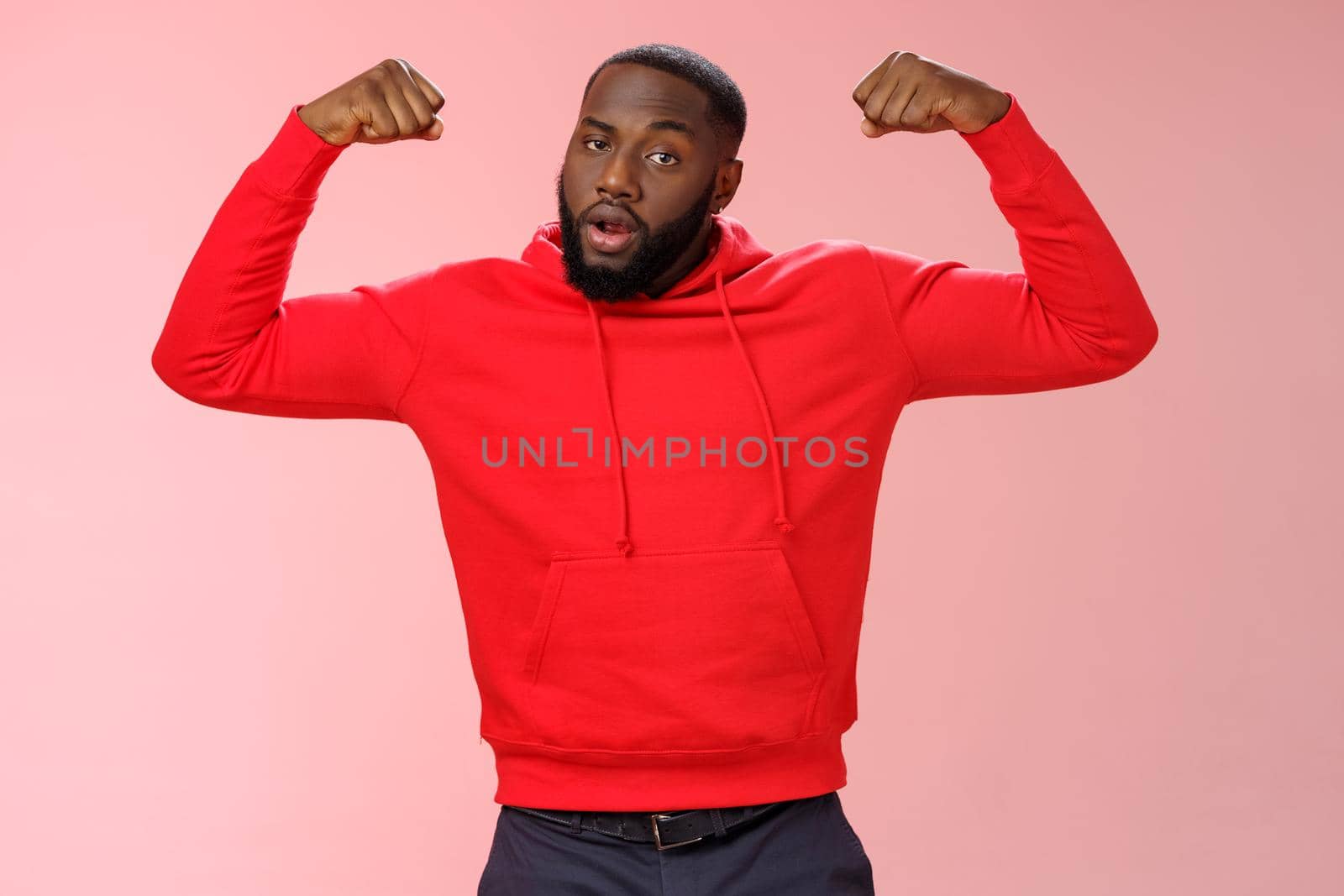 Strong cheeky serious-looking african american sportsman in red hoodie bragging showing-off raise hands excercising show big muscles biceps, standing confident pink background. Copy space