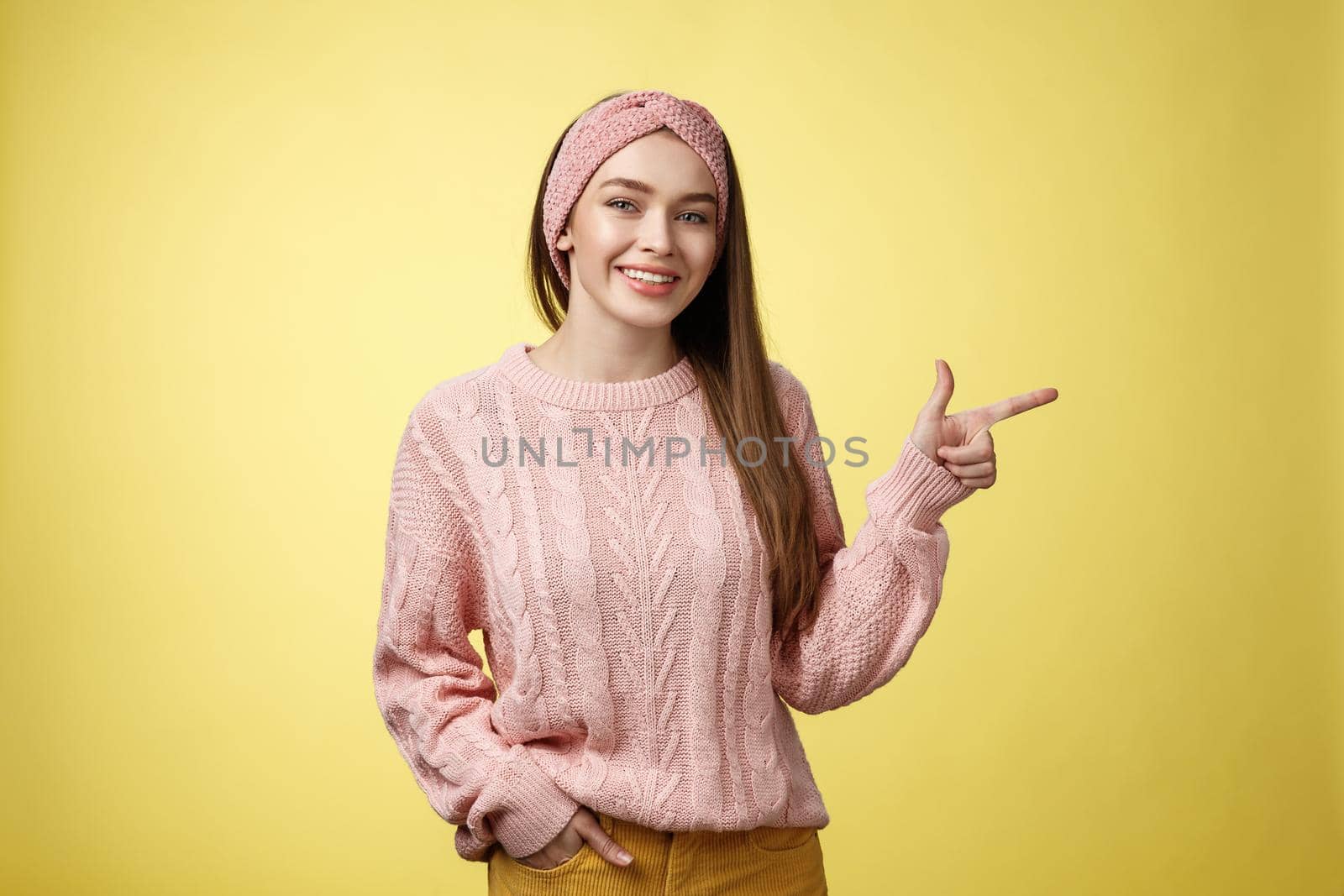 Lifestyle, people, pointing concept. Attractive young girl with long straight hair wearing sweater pointing aside using fore finger, indicating copy space smiling cheerful, positive over yellow wall by Benzoix