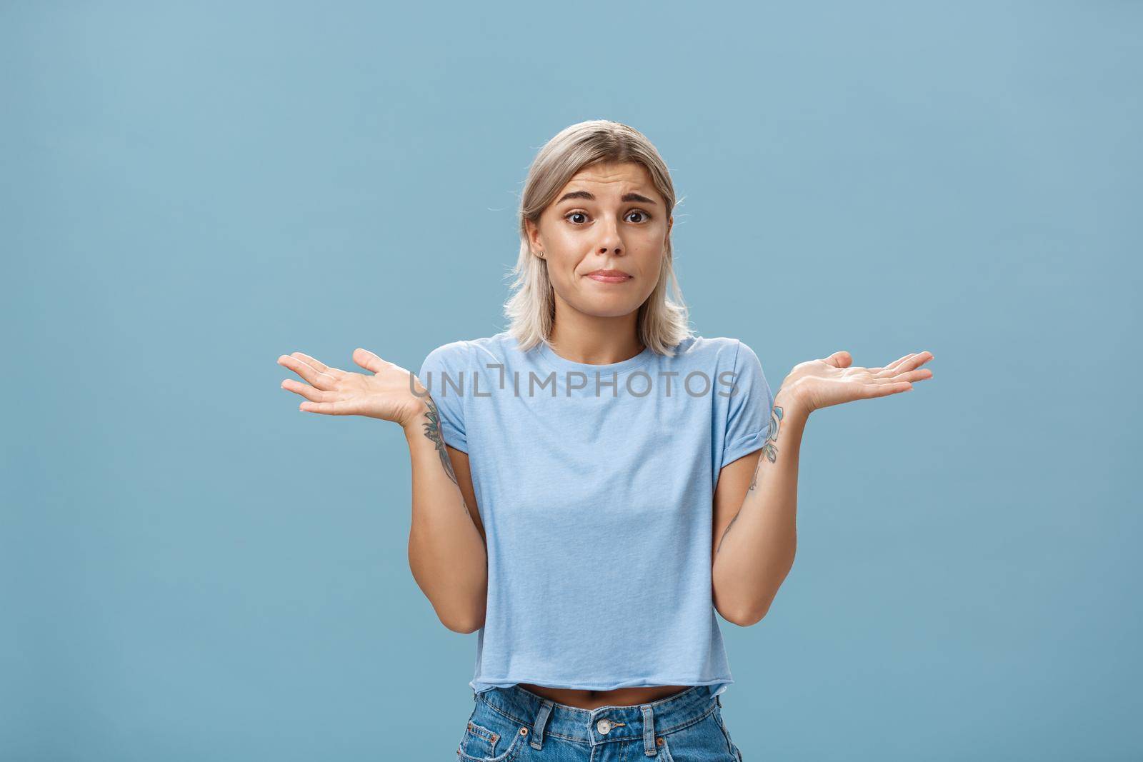No idea how help sorry. Portrait of unsure gloomy girlfriend feeling regret shrugging with hands near shoulders frowning and pursing lips being uncertain and unaware over blue background. Body language concept