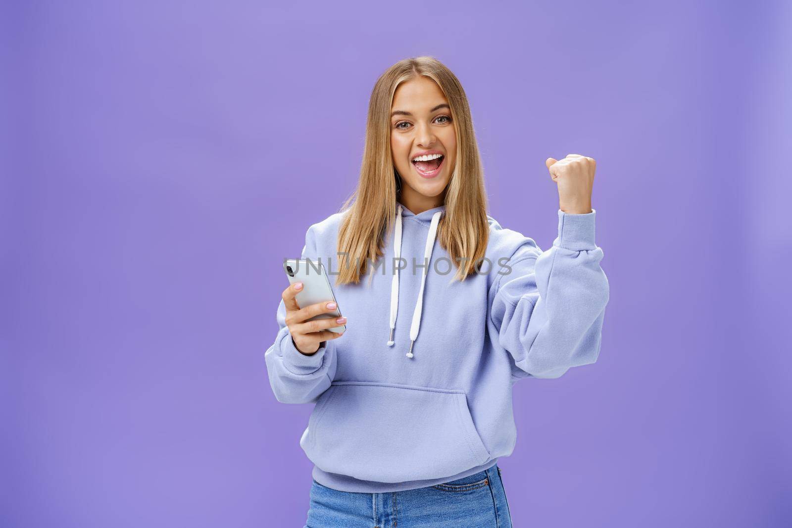 Yes we did it. Pleased excited woman in hoodie raising fist in celebration and success gesture smiling broadly triumphing holding smartphone reacting to positive news in cellphone over purple wall. Technology and emotions concept