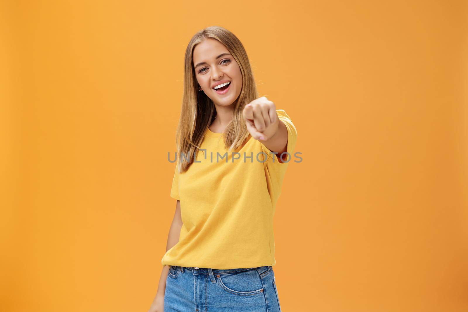 Woman directing at camera with index finger and smiling friendly as if talking to us standing amused and happy with confident carefree expression pulling arm towards to point over orange background. Lifestyle.