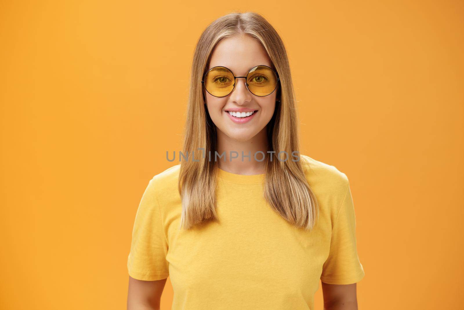 Stylish self-assured charming european woman with fair hair and tanned skin in yellow t-shirt and sunglasses smiling broadly amused and cheerful gazing at camera posing against orange background by Benzoix