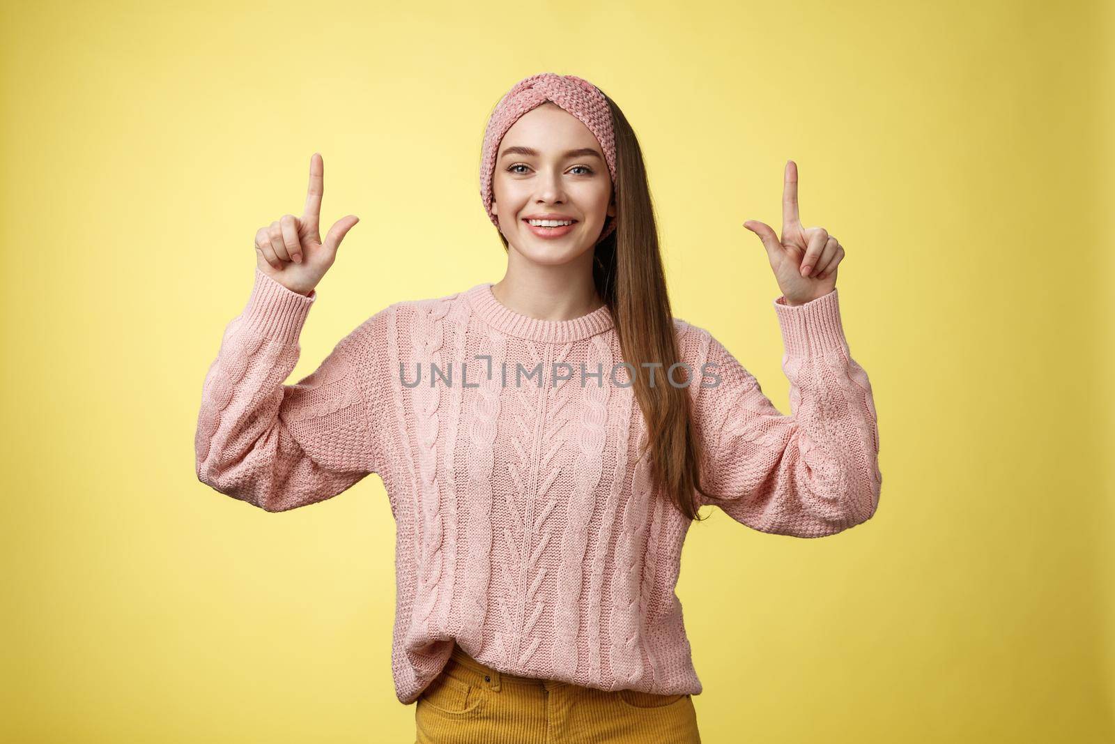 Easygoing beautiful young female student in knitted sweater, headband pointing up, promoting advertisement, smiling happily, feeling positive posing in good mood, grinning at camera over yellow wall.