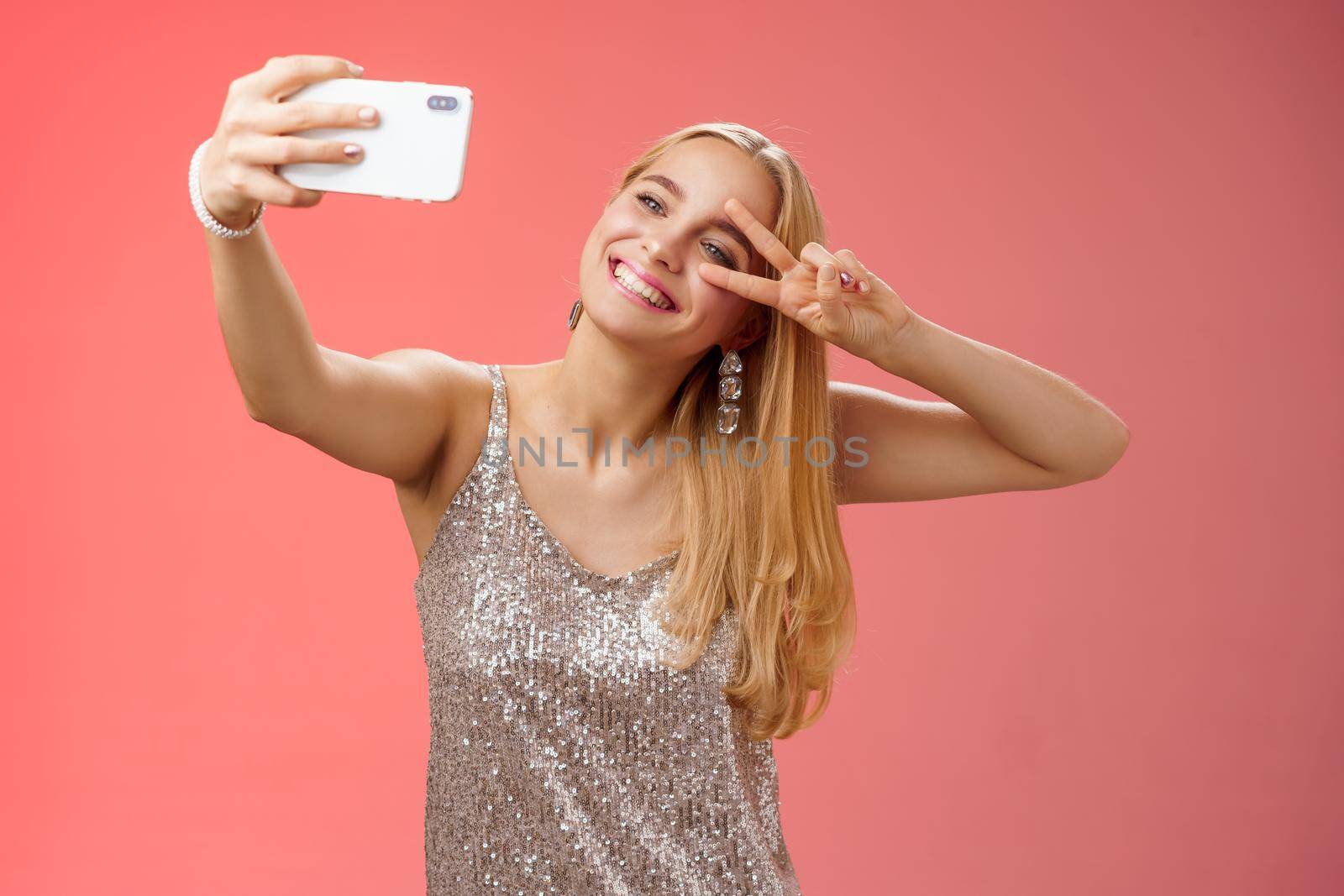Lifestyle. Stylish fabulous glamour young blond woman in glittering silver dress tilting head carefree show peace gesture victory sign extend arm holding smartphone taking selfie recording video post online.
