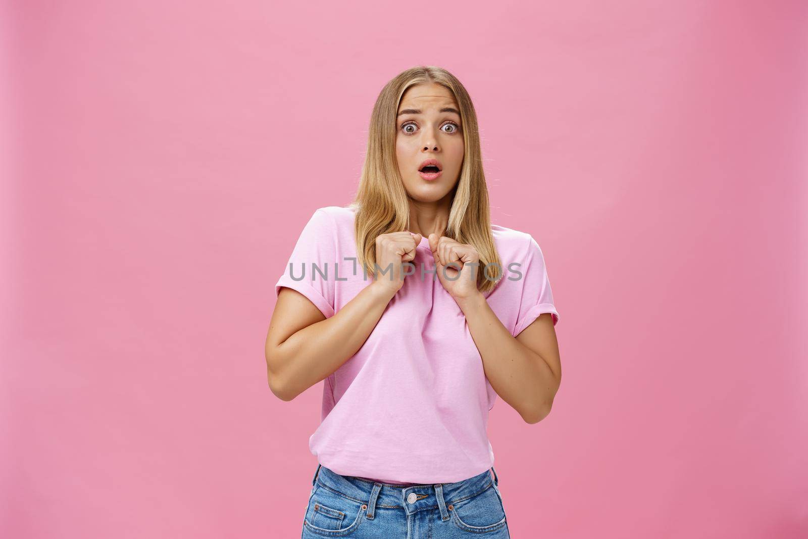 Woman gasping from fright being scared by someone pressing clenched hands to chest bending backwards looking worried and terrified at camera being shocked with unexpected bang over pink wall by Benzoix