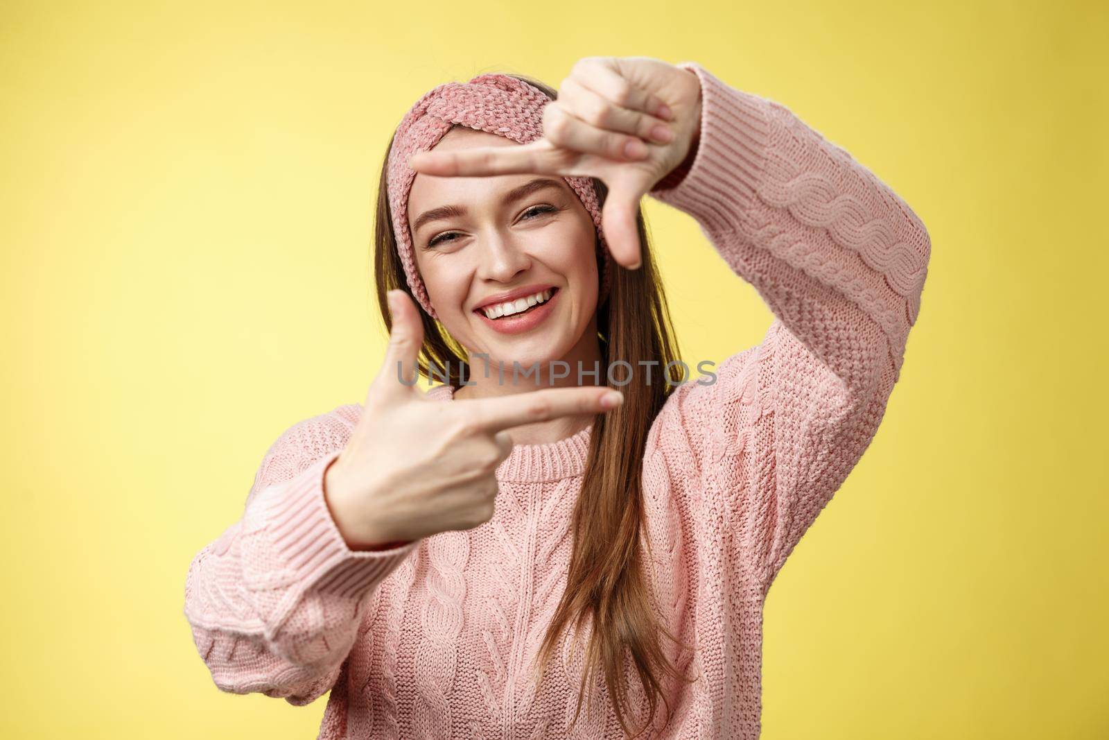 Charming outgoing, happy girl smiling cheerful, making frame of fingers looking through it excited and joyful, grinning, planning future, picturing how put furniture, posing cute over yellow wall.