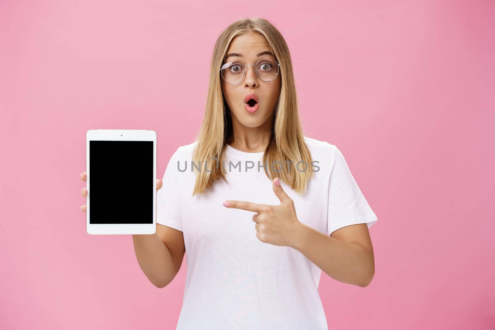 Shocked intelligent cute female in glasses and white t-shirt folding lips from amazement in wow sound gasping popping eyes amused holding digital tablet pointing at gadget screen over pink wall. Lifestyle.