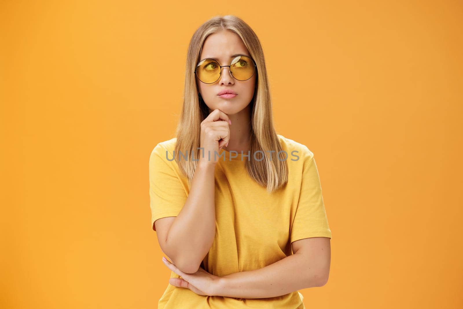 Troubled and concerned young stylish woman facing troublesome problem thinking holding hand on chin pursing lips looking at upper right corner thoughtfully standing against orange background. Lifestyle.