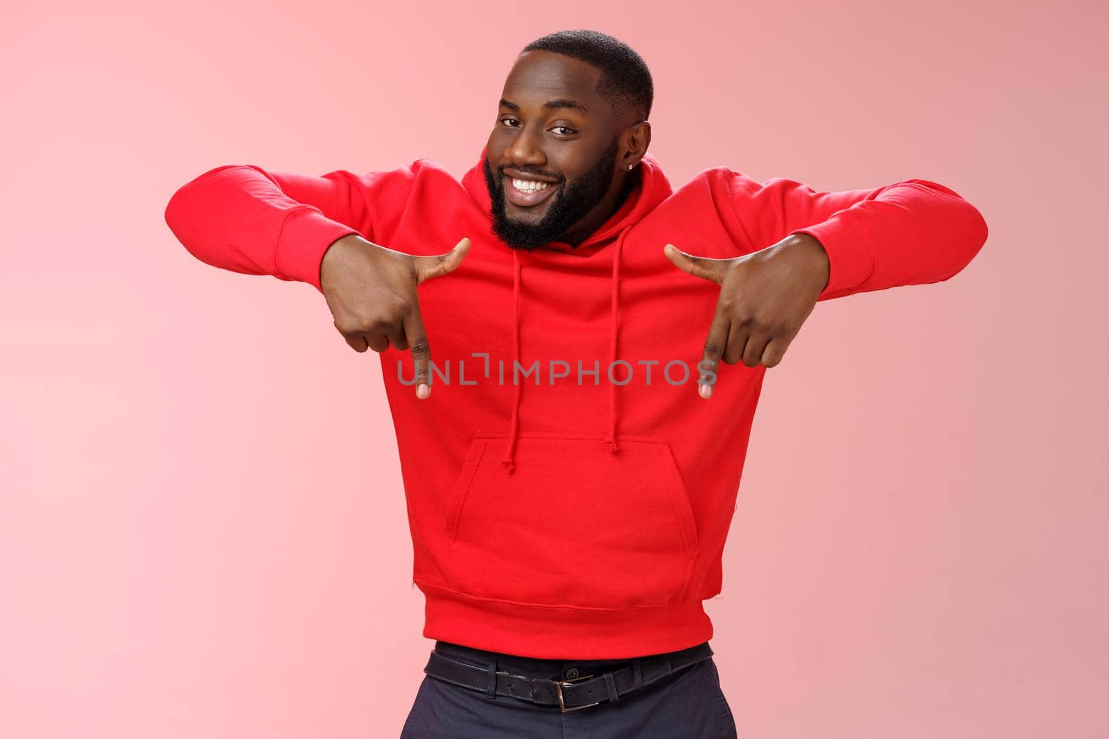 Sassy young charismatic handsome bearded black guy in red hoodie pointing down index fingers smiling broadly showing awesome copy space solution cool product for you, standing pink background.