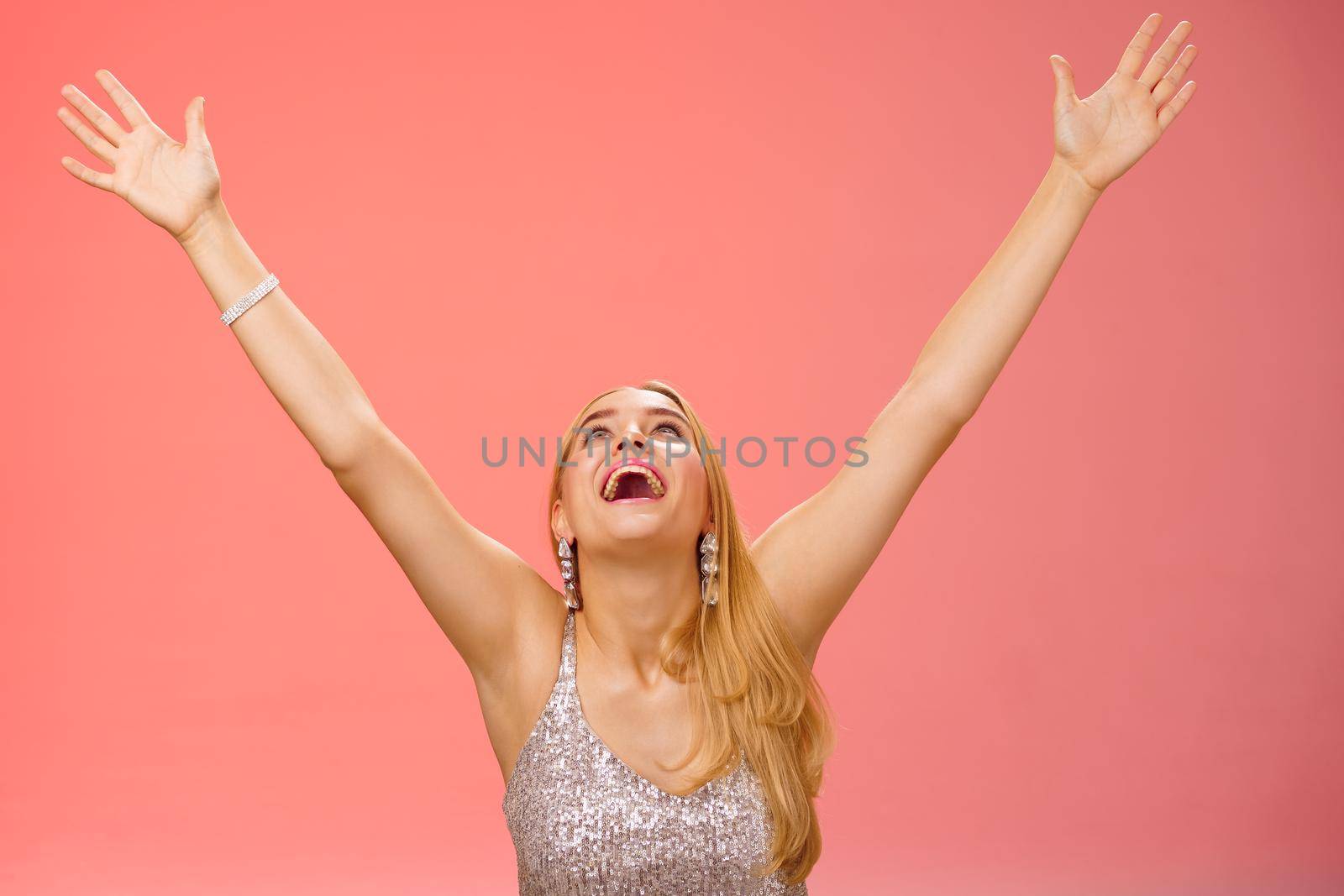 Happy emotive smiling overwhelmed young blond woman in silver dress raise hands sky thank god joyfully signed contract got job rejoicing red background celebrating victory good news, triumphing.