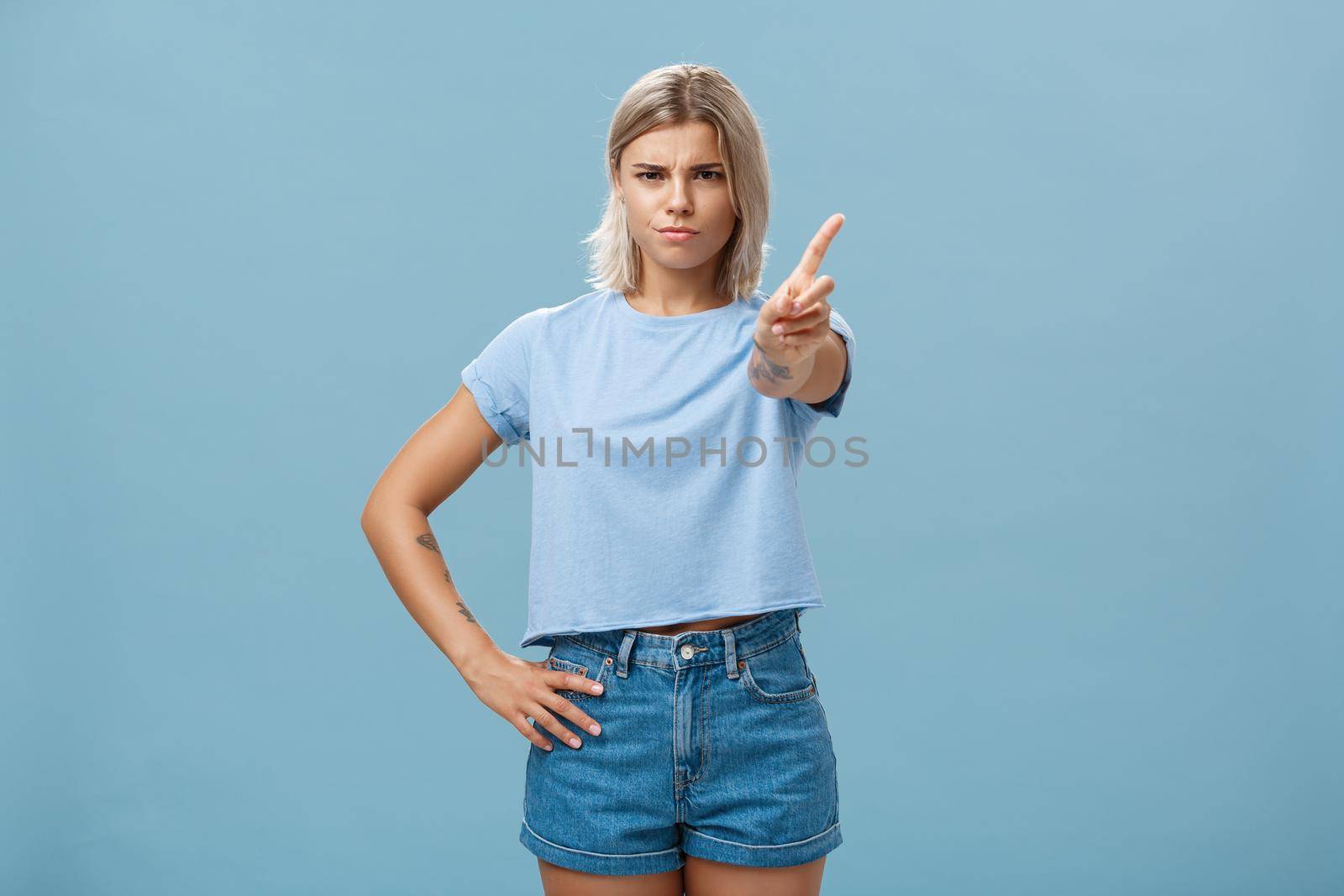 You better not do it. Serious-looking displeased strict elder sister with fair hair and tanned skin frowning holding hand on waist shaking index finger in forbid or stop gesture over blue background by Benzoix