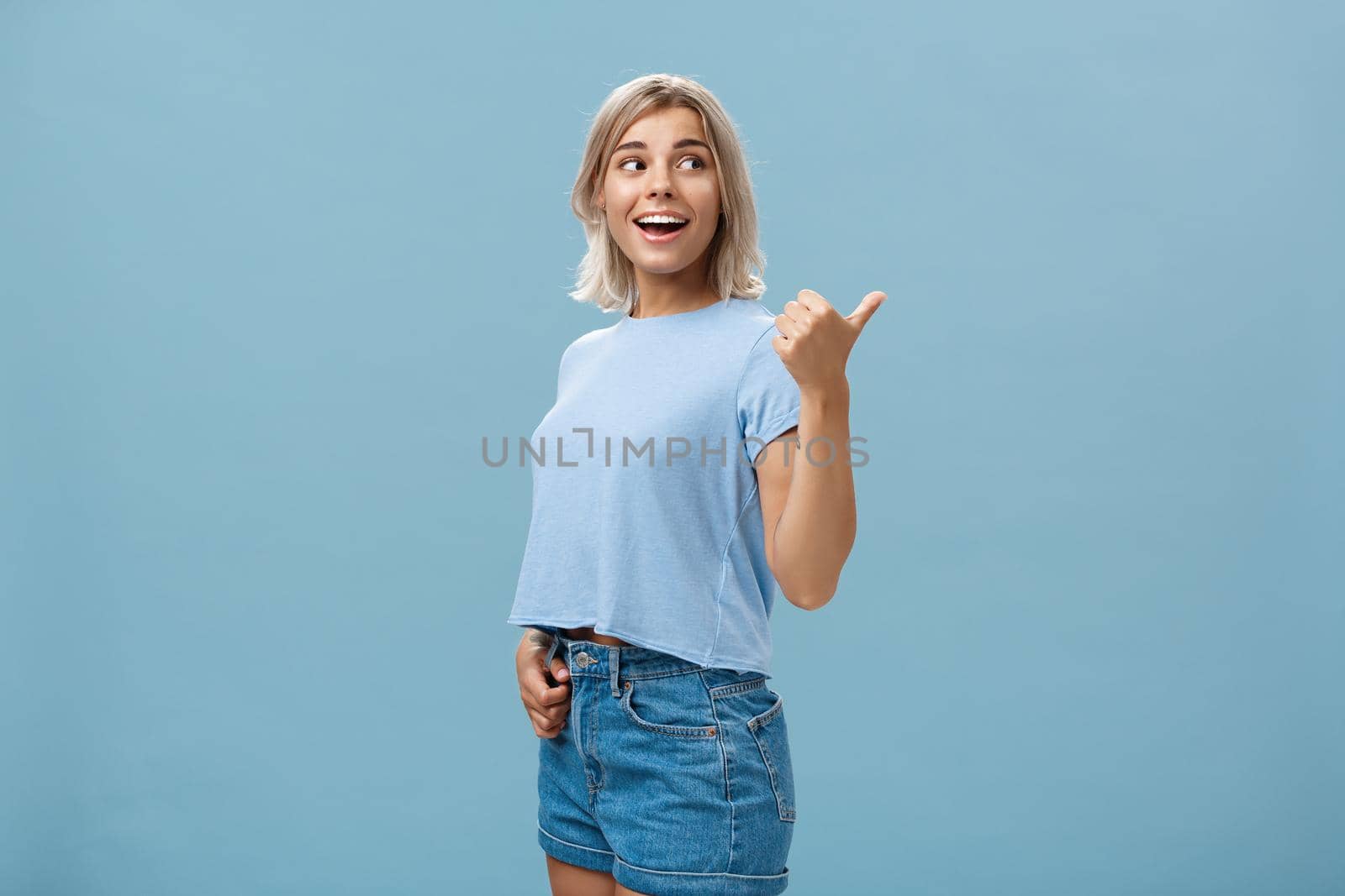 Charming sociable and friendly stylish female with blond hair turning right to show way or while discussing something talking with relaxed carefree look posing over blue background in trendy outfit by Benzoix
