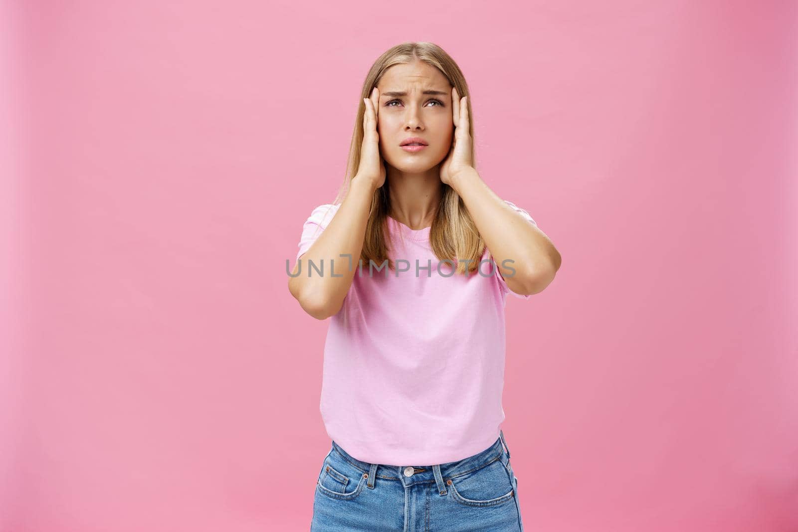 Woman trying recall important number touching temples with hands looking up concerned and focused having trouble to remember information standing intense against pink background by Benzoix