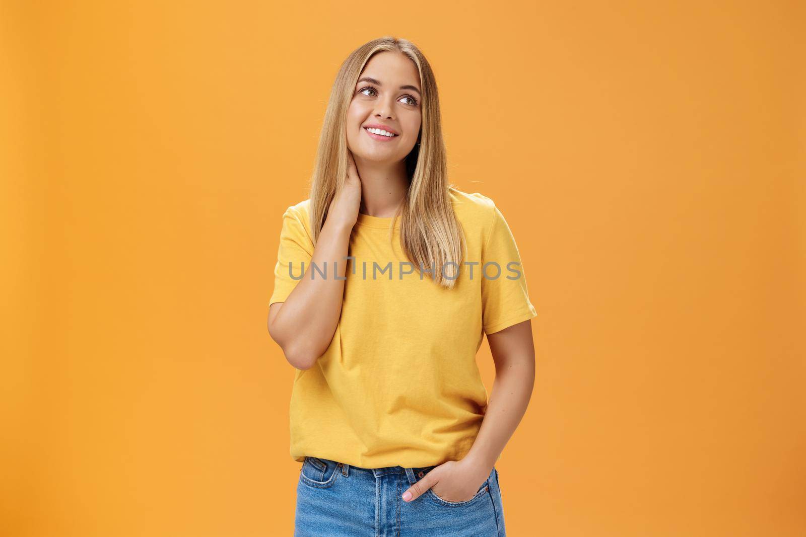 Pretty and tender cute european female independent freelancer in yellow t-shirt sighing touching neck and gazing dreamy at upper right corner with pleasant smile, posing over orange background by Benzoix