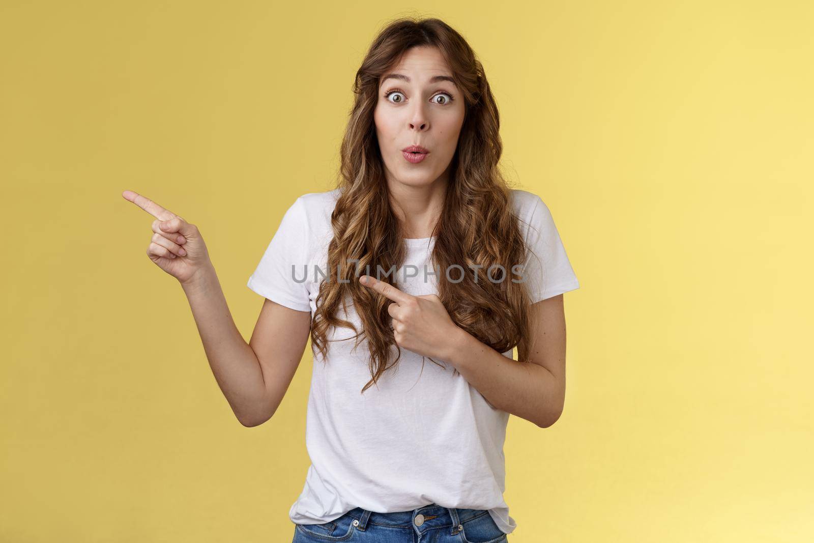 Awesome lets go look closer. Impressed surprised attractive caucasian curly-haired girlfriend fold lips admiration tempting try new product pointing index fingers left stare camera astonished excited by Benzoix
