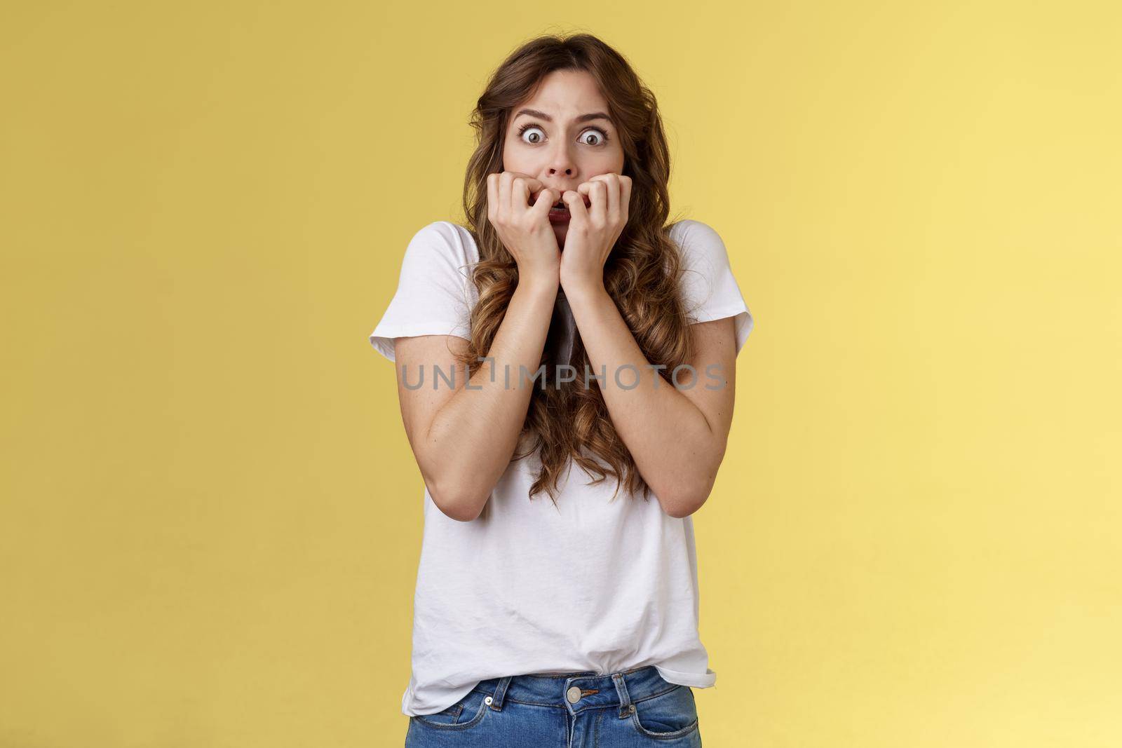 Shocked speechless scared stunned curly-haired caucasian woman gasping hold fingers mouth biting nails stare camera frightened express fear terrified scary movie stand yellow background.