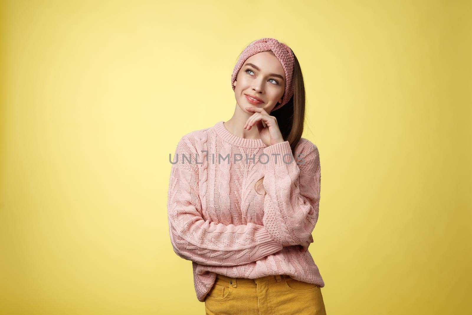 Dreamy attractive and stylish schoolgirl in knitted warm sweater, headband, thinking as smiling satisfied and devious having idea, looking at upper left corner thoughtful, daydreaming, planning.