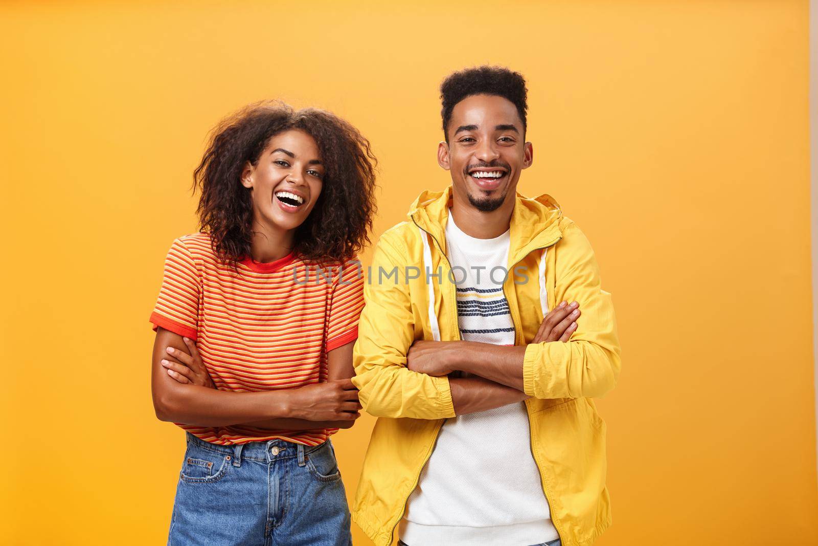 Two african american man and woman being best friends laughing out loud watching funny movie in cinema all dressed up in stylish outfit standing with hands crossed on chest and amused expression by Benzoix