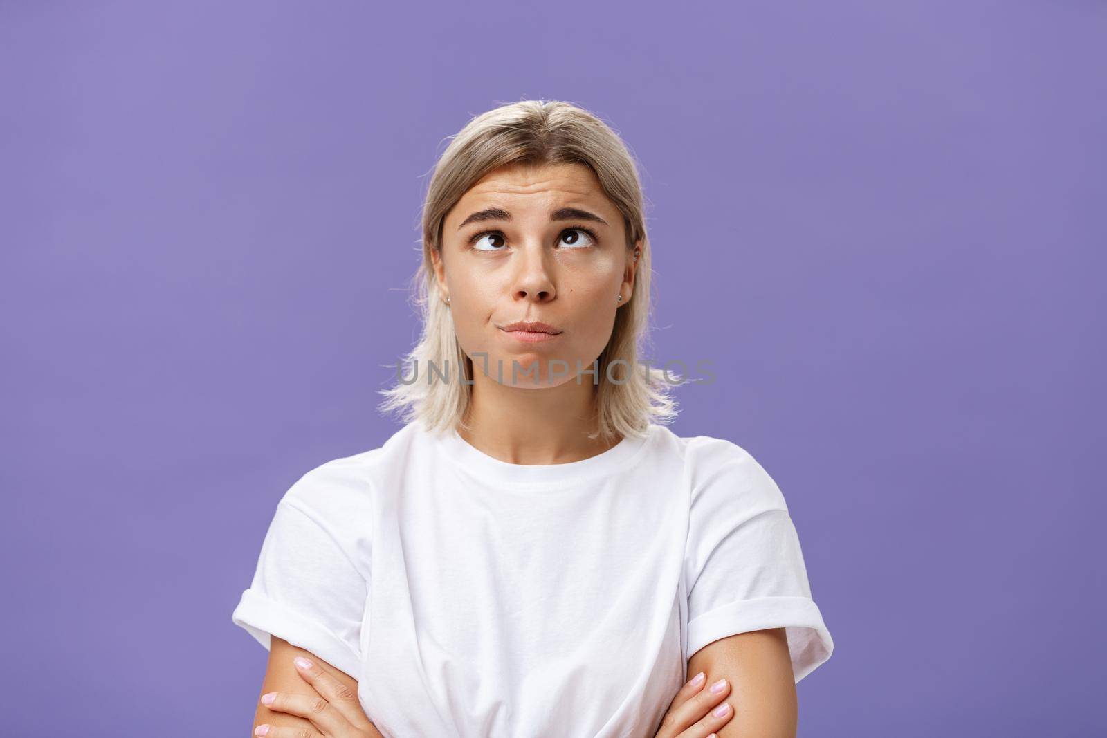 Mind going crazy. Portrait of funny tired attractive young woman with fair hair and tanned skin squinting rolling eyes aside and looking up fooling around over purple background from boredom. Emotions concept