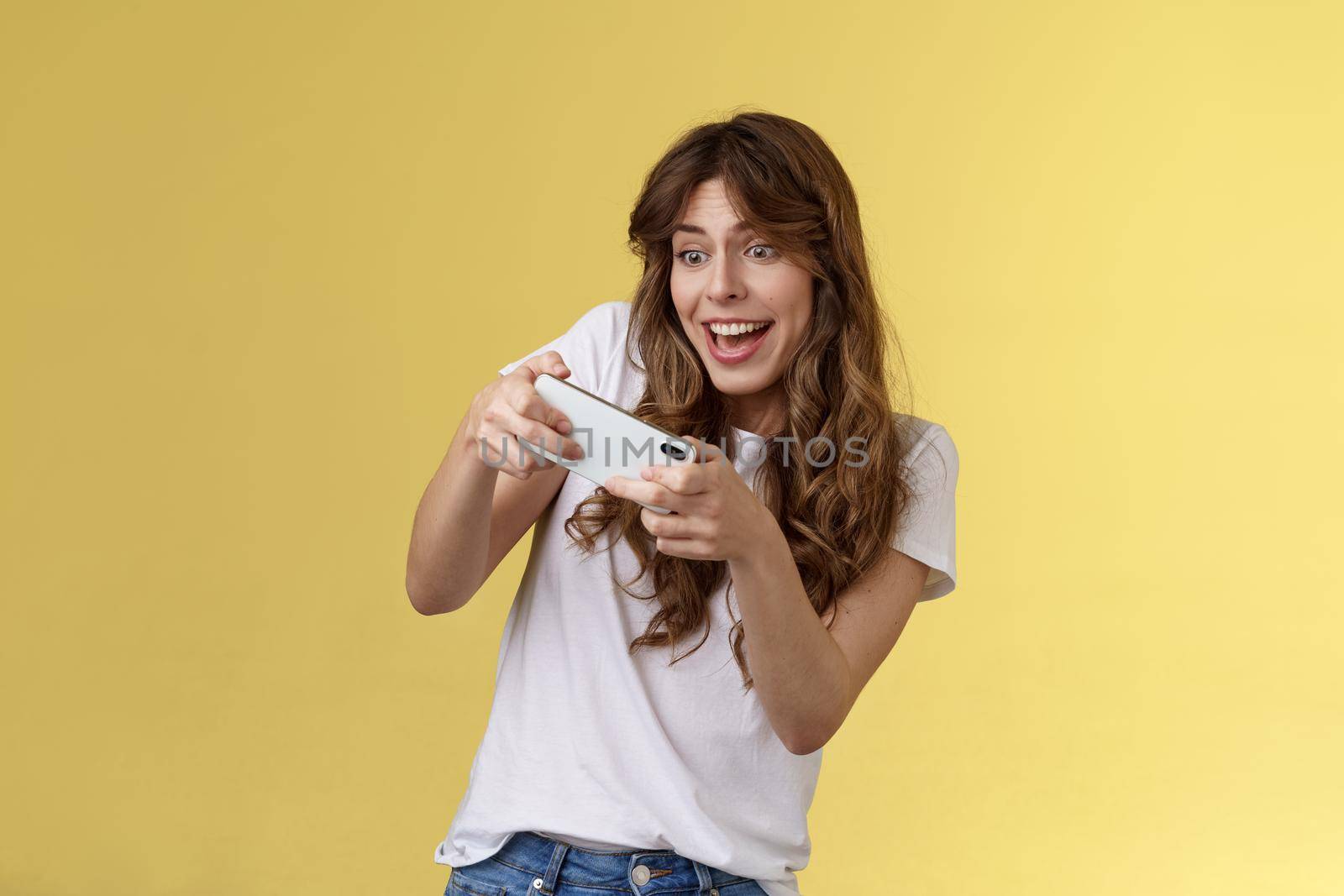 Excited playful enthusiastic girl tilting sideways playing awesome interesting smartphone game car racing smiling determined focuse gaming hold mobile phone horizontal tap display yellow background by Benzoix