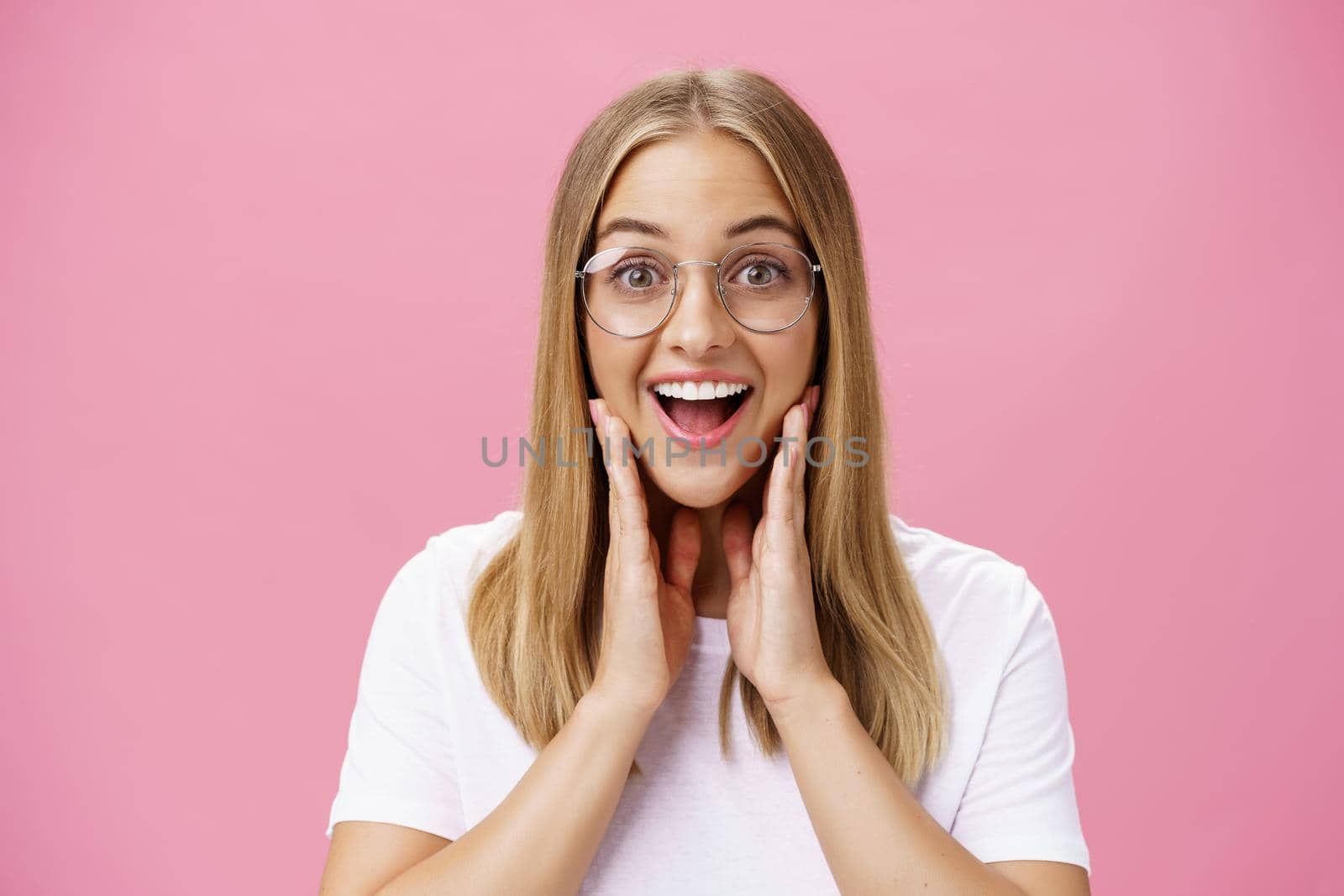 Woman liking new glasses picked in store feeling happy finding right frame touching cheeks from happiness smiling cheerfully at camera wearing transparent eyewear and t-shirt over pink wall by Benzoix