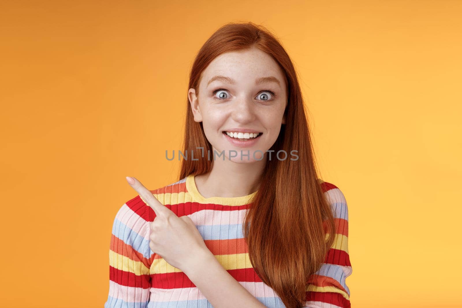 Excited thrilled good-looking young silly redhead girl surprised pop eyes camera gasping impressed pointing upper left corner see super star grinning excitement delight, orange background. Copy space