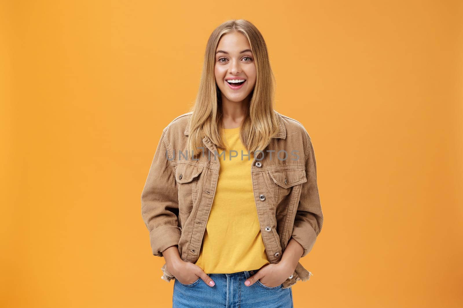 Charismatic tanned woman in corduroy jacket and yellow t-shirt ready for chilly autumn walk with friends smiling joyfully gazing entertained at camera holding hand in pockets casually over orange wall. Lifestyle.