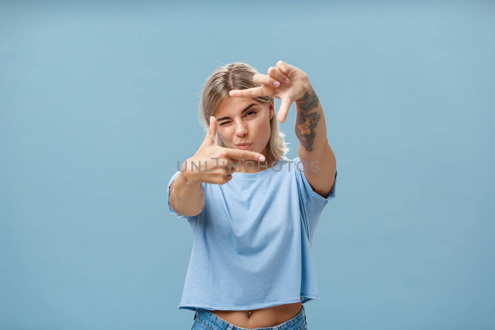 Artistic and creative female designer in stylish t-shirt closing one eye folding lip and making frame gesture while looking through it as if taking measurement or picturing something over blue wall. Copy space