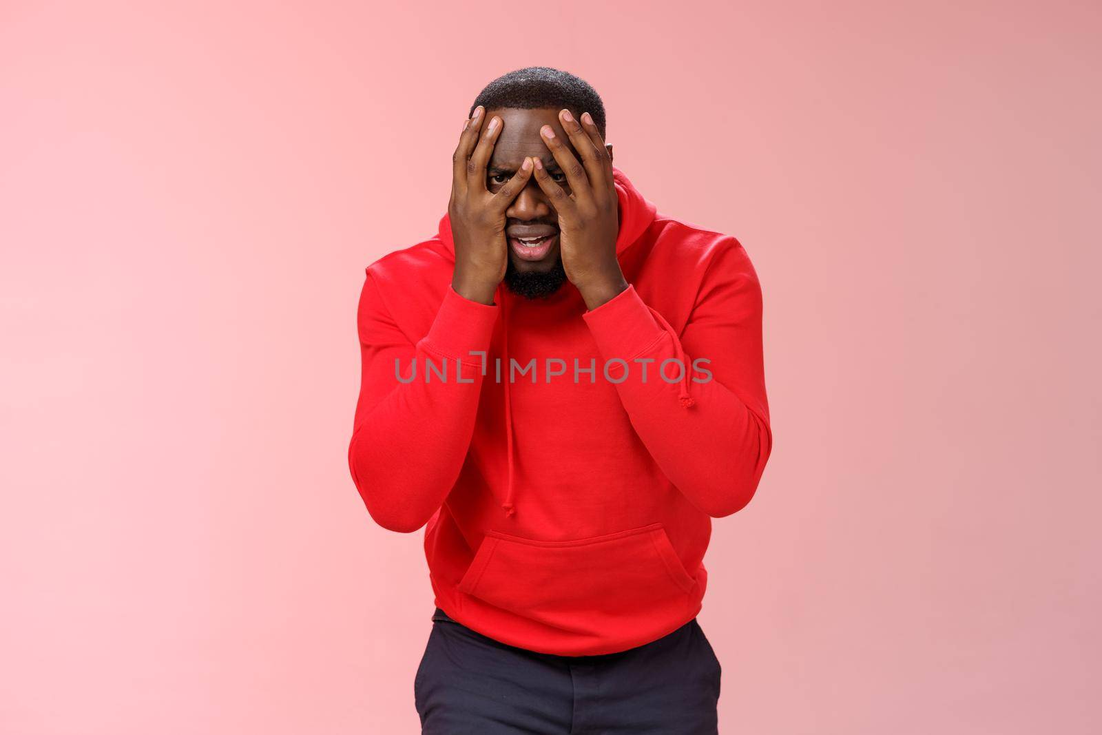 Shocked upset young african american bearded guy in red hoodie facepalm hide face hands peeking through fingers sad devastated lose hope, standing sad pink background grieving feel sorrow.