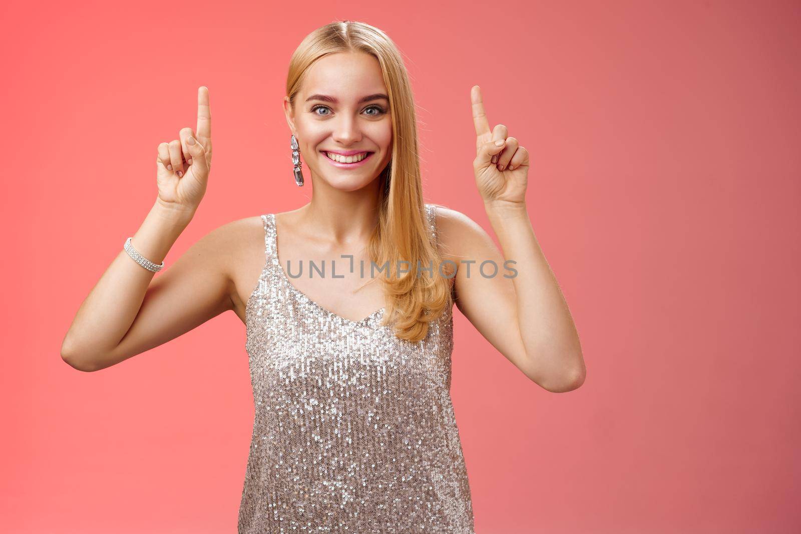 Excited gorgeous blond european woman in silver shiny elegant dress raise hands pointing up showing impressive incredible advertisement smiling happily thrilled wanna take closer look, red background.