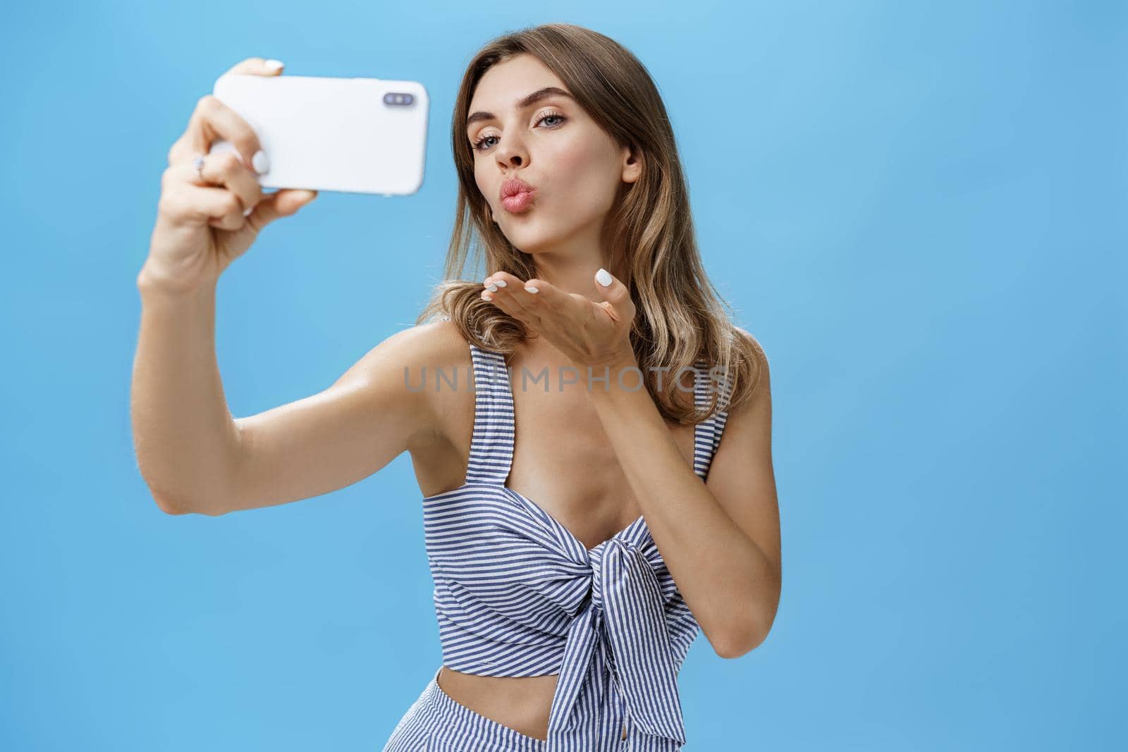 Outgoing and self-assured glamorous woman like taking pictures of herself holding smartphone making sensual and flirty selfie folding lips raising palm to send wind kiss at screen loving followers. Technology, lifestyle, social network concept