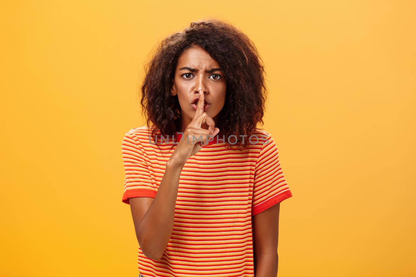 Shh do not disturb. Portrait of serious-looking bossy and displeased cute african american girl with afro hairstyle frowning shushing at camera with index finger over mouth forbidding speak loud.
