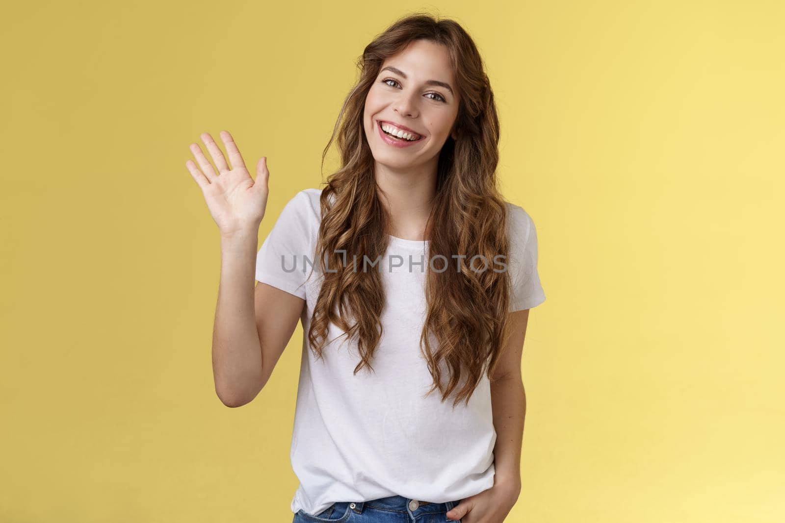 Modest cute friendly outgoing attractive european woman long curly haircut waving palm sociable conversation introduce herself smiling broadly say hi hello greeting coworker yellow background by Benzoix