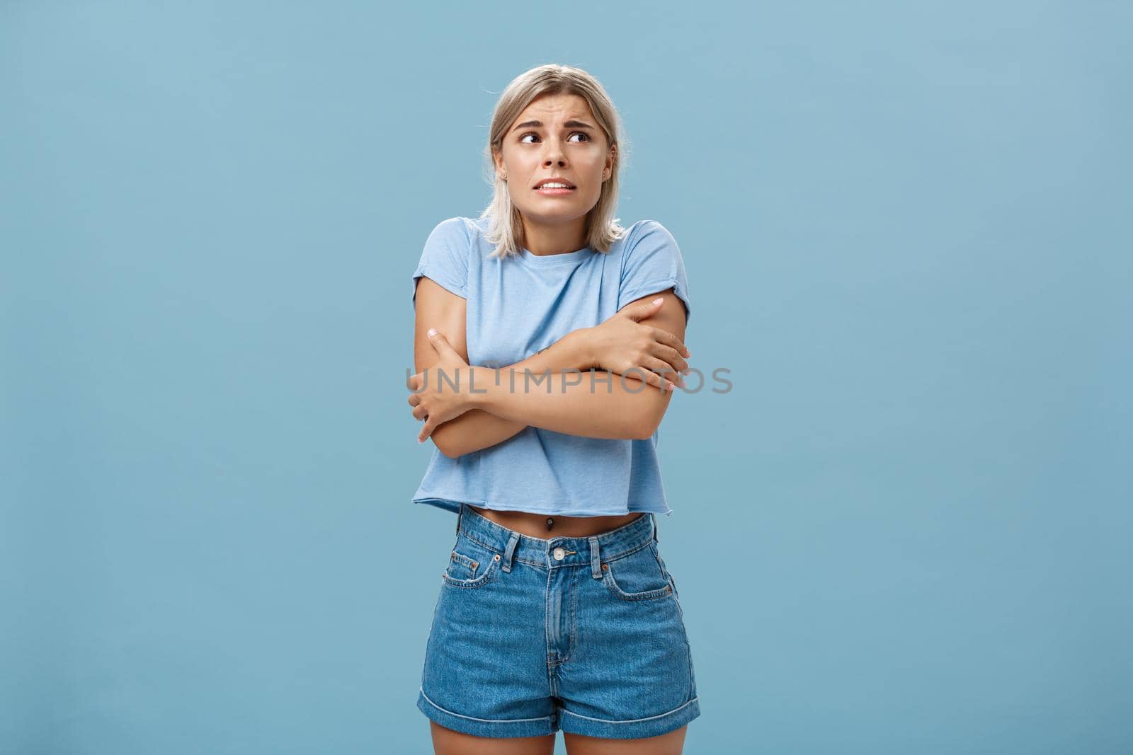 Turn off air-cooler I am freezing in summer. Displeased cute caucasian blonde girl in trendy t-shirt hugging herself while trembling from cold shaking gazing right with worried look over blue wall.