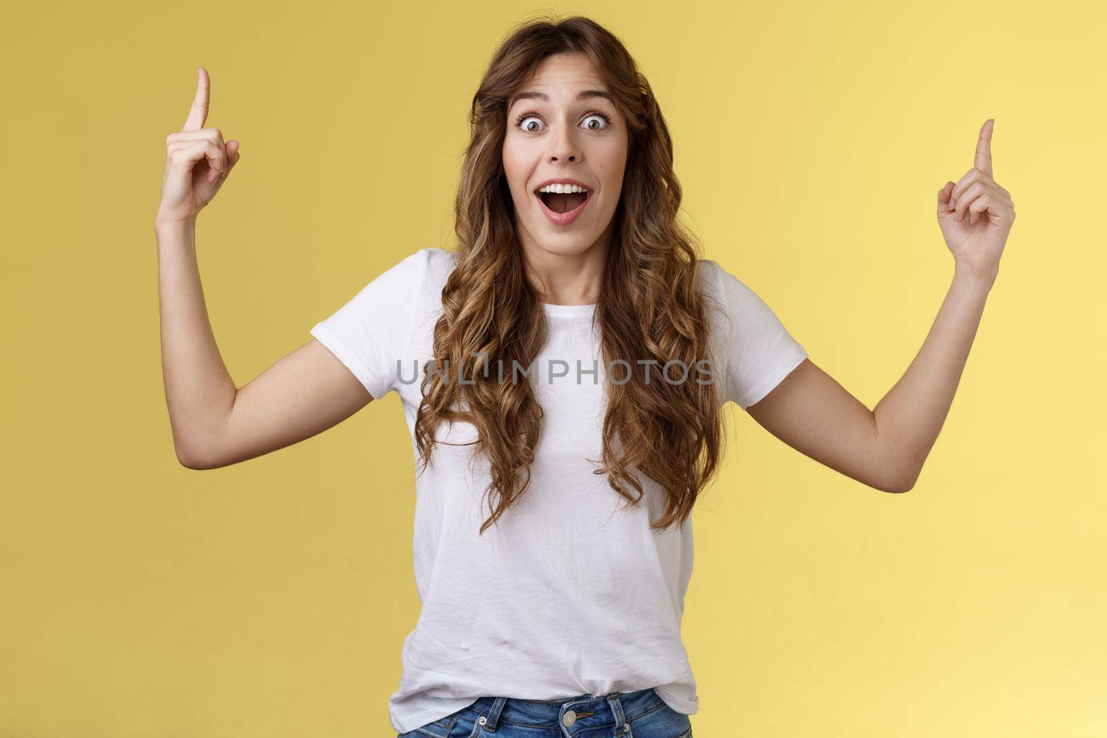 Fascinated speechless excited attractive curly-haired girl impressed stare camera surprised glad appreciation awesome gift receive incredible offer pointing up index fingers yellow background.