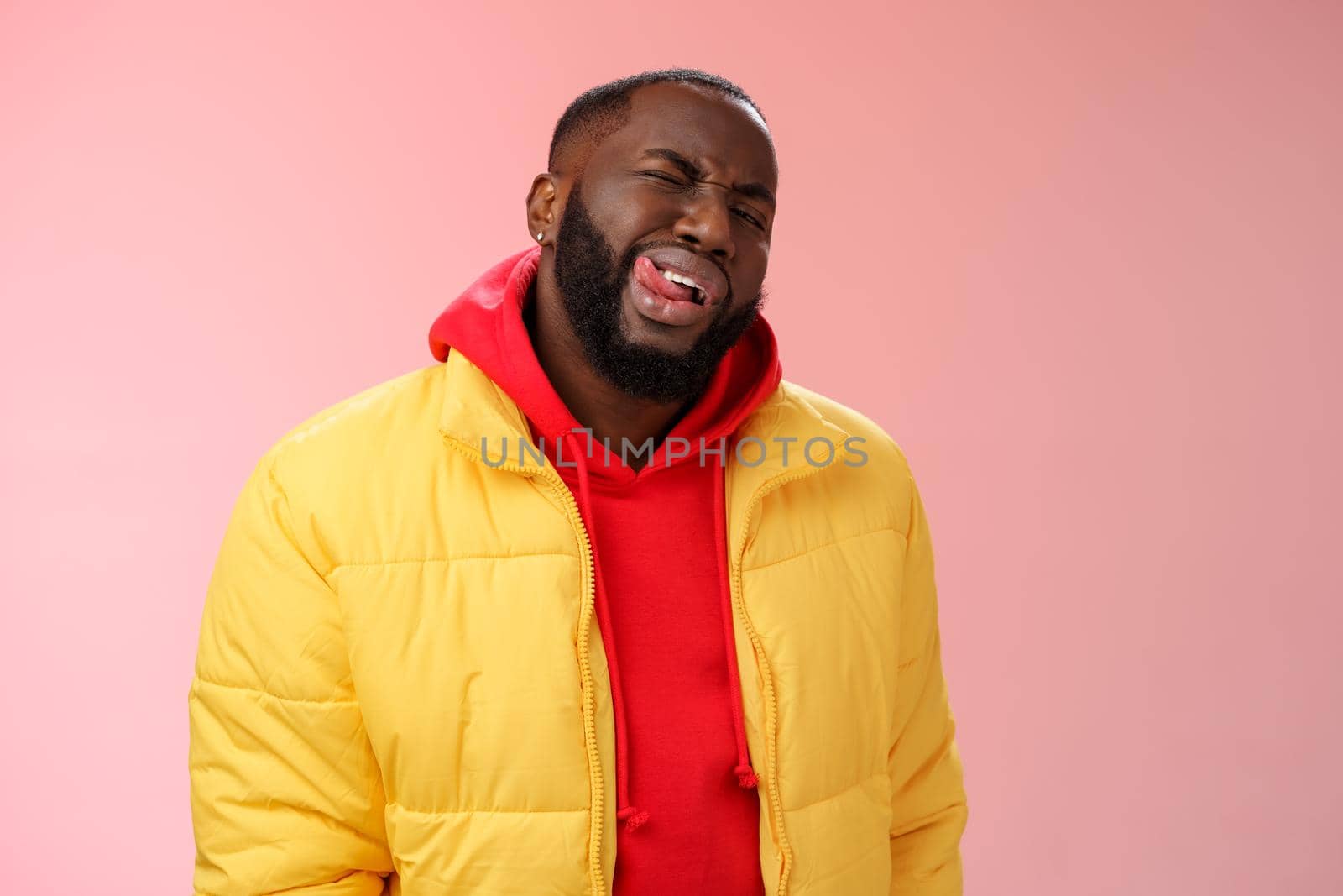 Cheeky stylish funny african bearded man in yellow trendy jacket red hoodie show tongue sassy flirty gaze winking camera trying impress woman pretend macho, standing pink background.