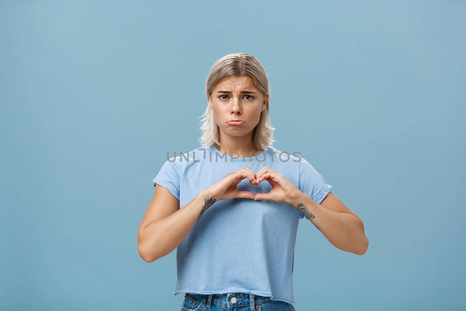 Heart being broken. Sad and gloomy heartbroken girl with blond hair tattoos on arms and tanned skin pursing lips whining and complaining making love sign over breast standing unhappy near blue wall.