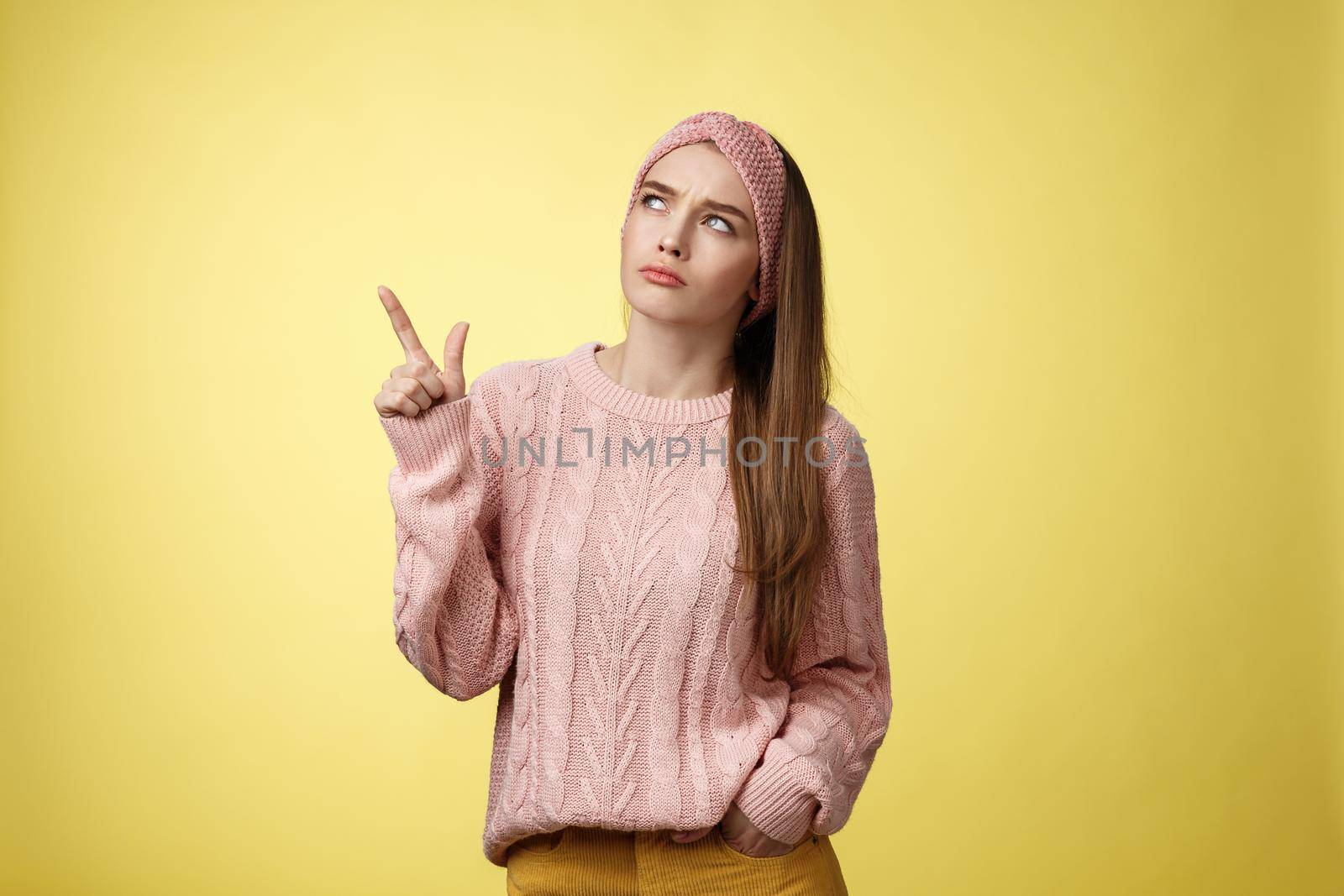 Frustrated upset, gloomy cute young woman frowning making disappointed grimace pointing, looking upper left corner displeased unsatisfied and unhappy posing confused against yellow background.