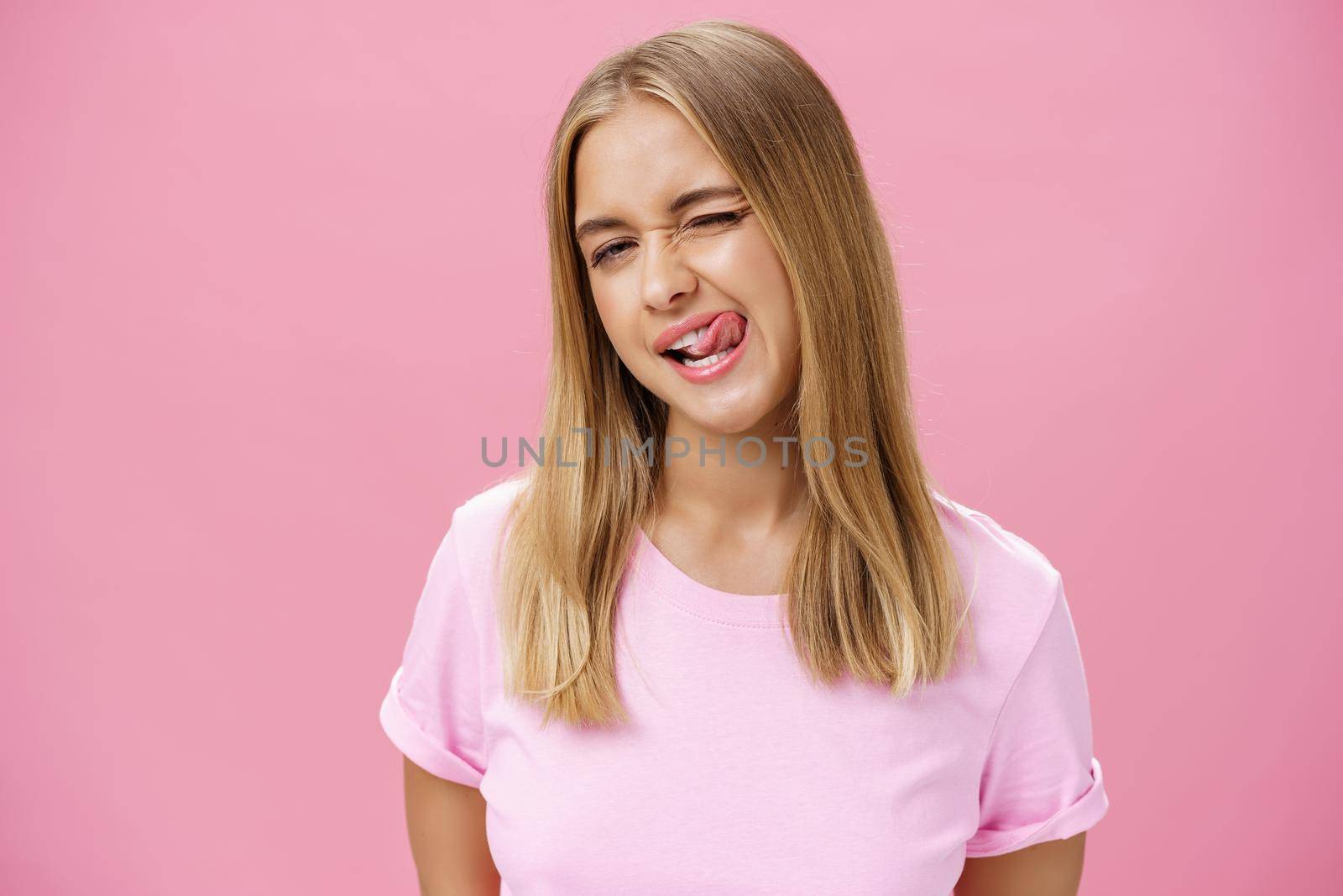 Playful flirty attractive young girl with fair straight hair in t-shirt winking at camera smiling and licking upper lip with tongue feeling enthusiastic and carefree against pink background. Emotions concept