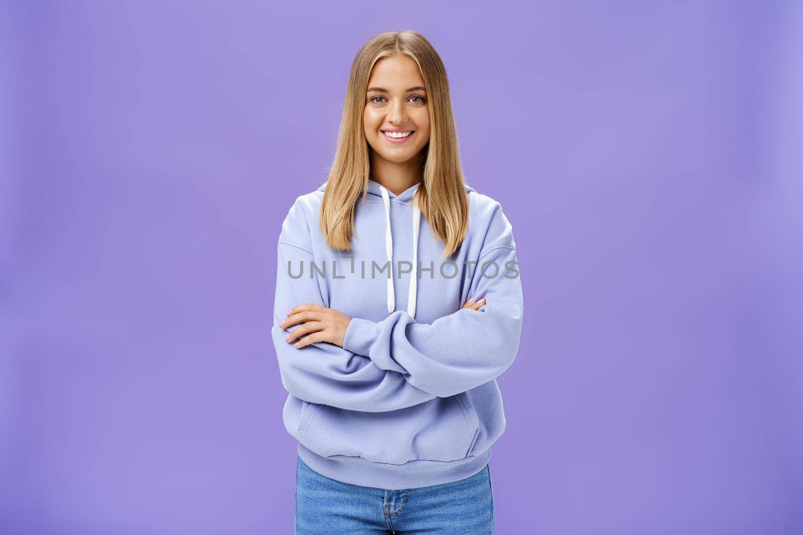 Portrait of charming charismatic european woman with tanned skin in trendy over-sized hoodie crossing hands against chest smiling broadly at camera standing self-assured against purple background. Lifestyle.